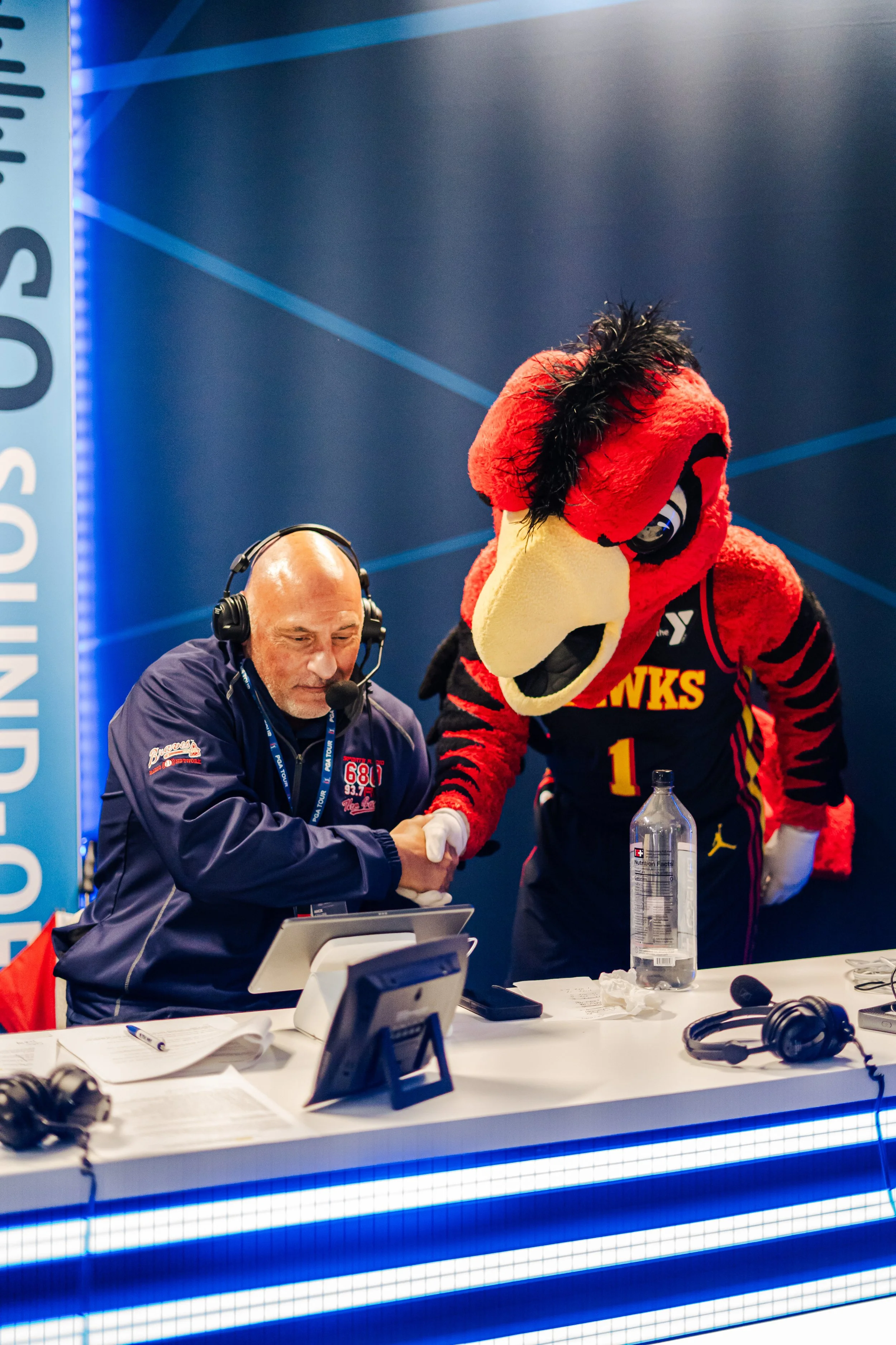 A radio broadcaster in a dark blue jacket shaking hands with a mascot dressed as a hawk wearing a sports jersey, sitting at a broadcasting desk with headphones, microphones, and papers.