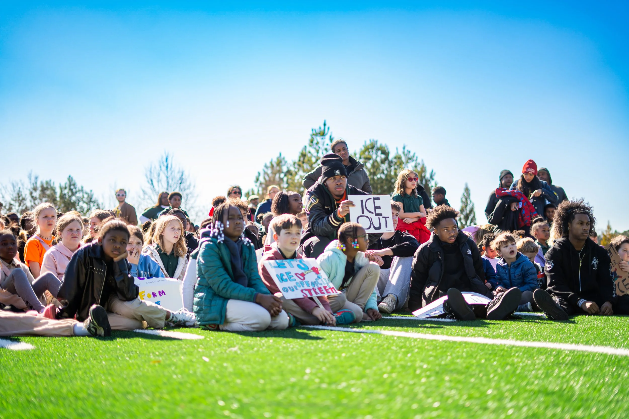 Children and teenagers sitting on a grassy field during a protest or rally, holding signs that advocate for climate or environmental action. The sky is clear and blue, with trees in the background.