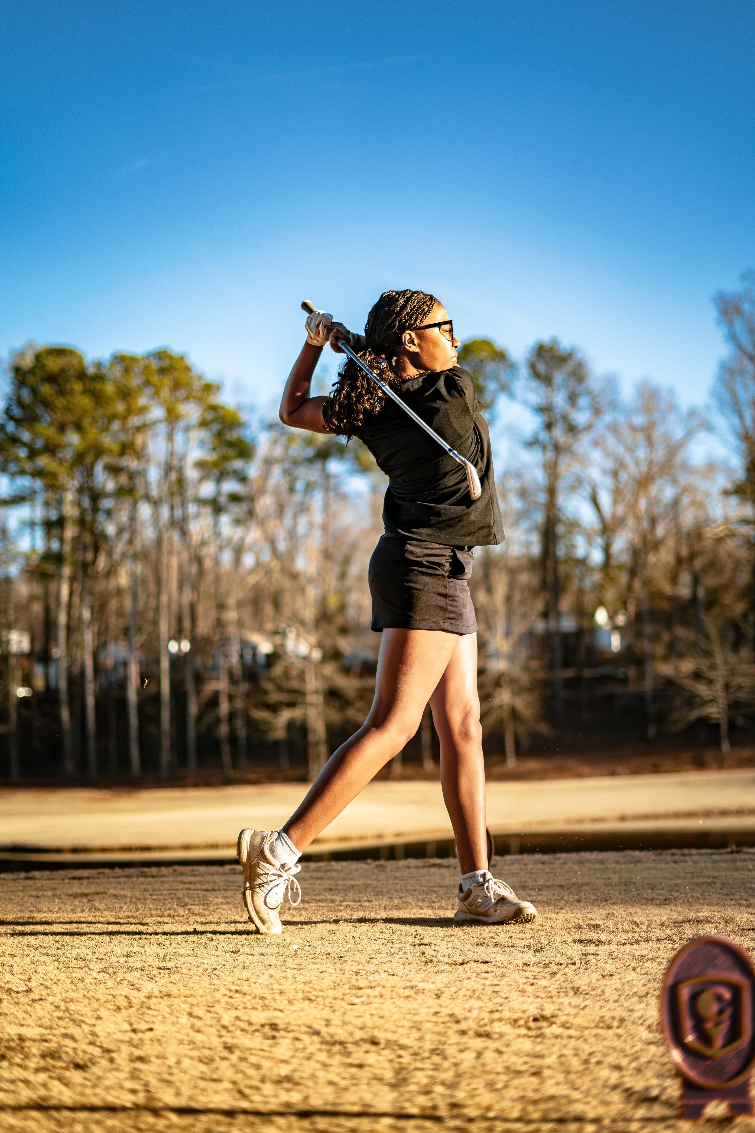 A young woman in black athletic clothing, wearing sunglasses and gloves, is swinging a golf club on a golf course under a clear blue sky with trees in the background.