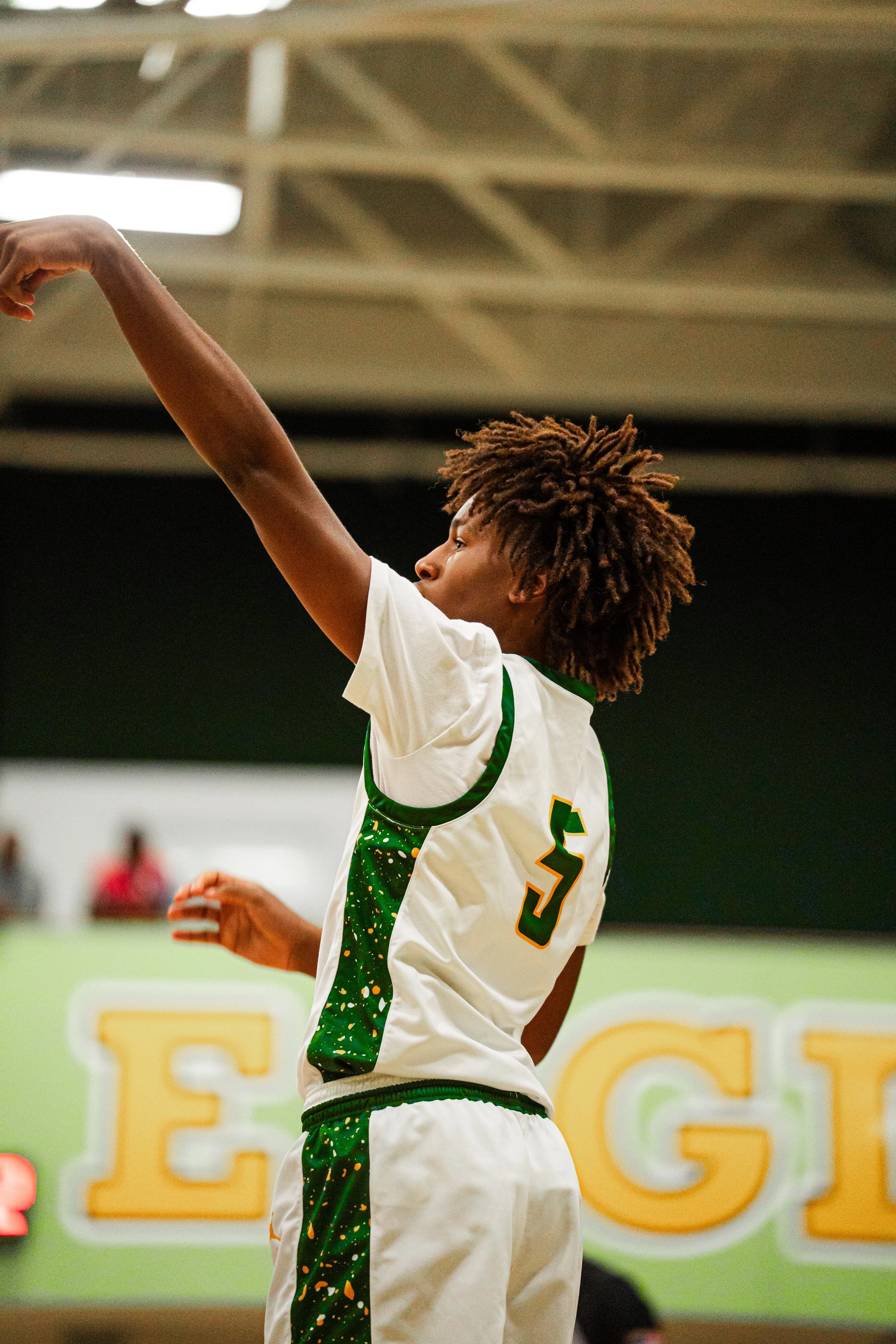 A young male basketball player wearing a white and green uniform with the number 5, shooting a basketball in an indoor gym.