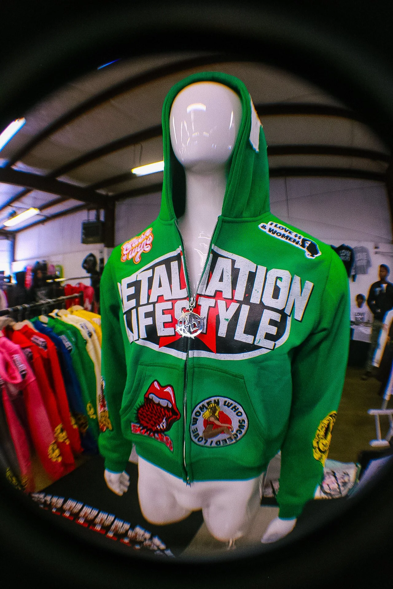 A mannequin wearing a bright green hoodie with various patches and graphic text, displayed in a retail store with colorful clothing in the background.