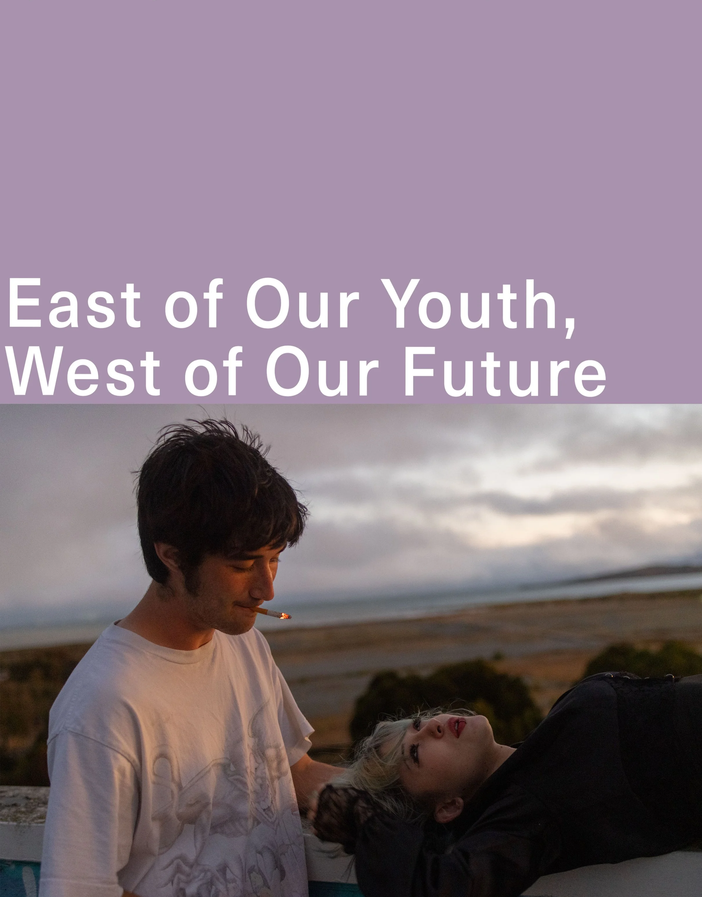 Two young people, one male and one female, at a beach at sunset. The male has dark hair and is smoking a cigarette, while the female has blonde hair and is lying on her side, looking up at him. There is a cloudy sky in the background.