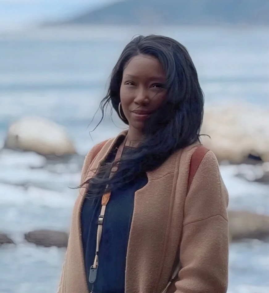 Westeigh, co-founder of Frame 1A, standing near a body of water with rocks in the background, she is wearing a tan sweater and a black top.