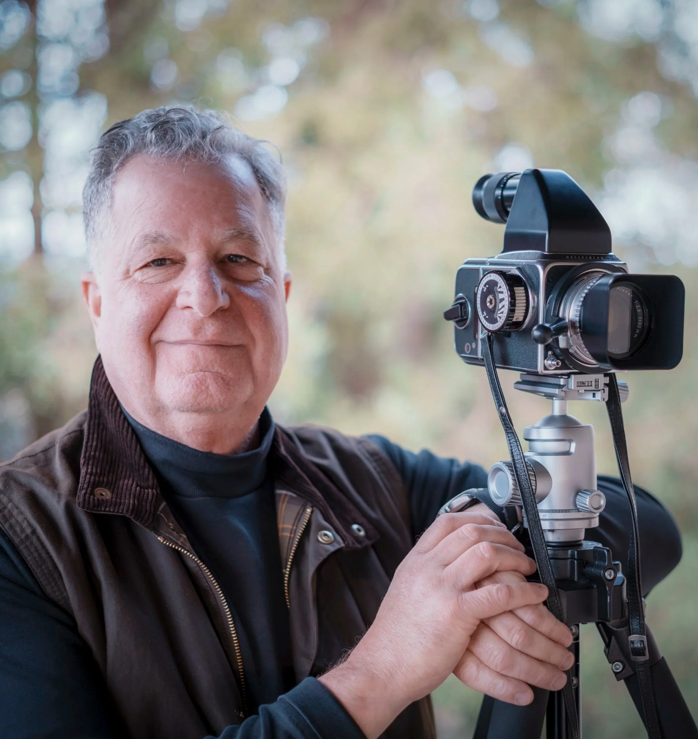 Jeff Heyman, co-founder of Frame 1A, with gray hair and fair skin standing by a Hasselblad camera outdoors, with a blurred background of trees and sunlight.