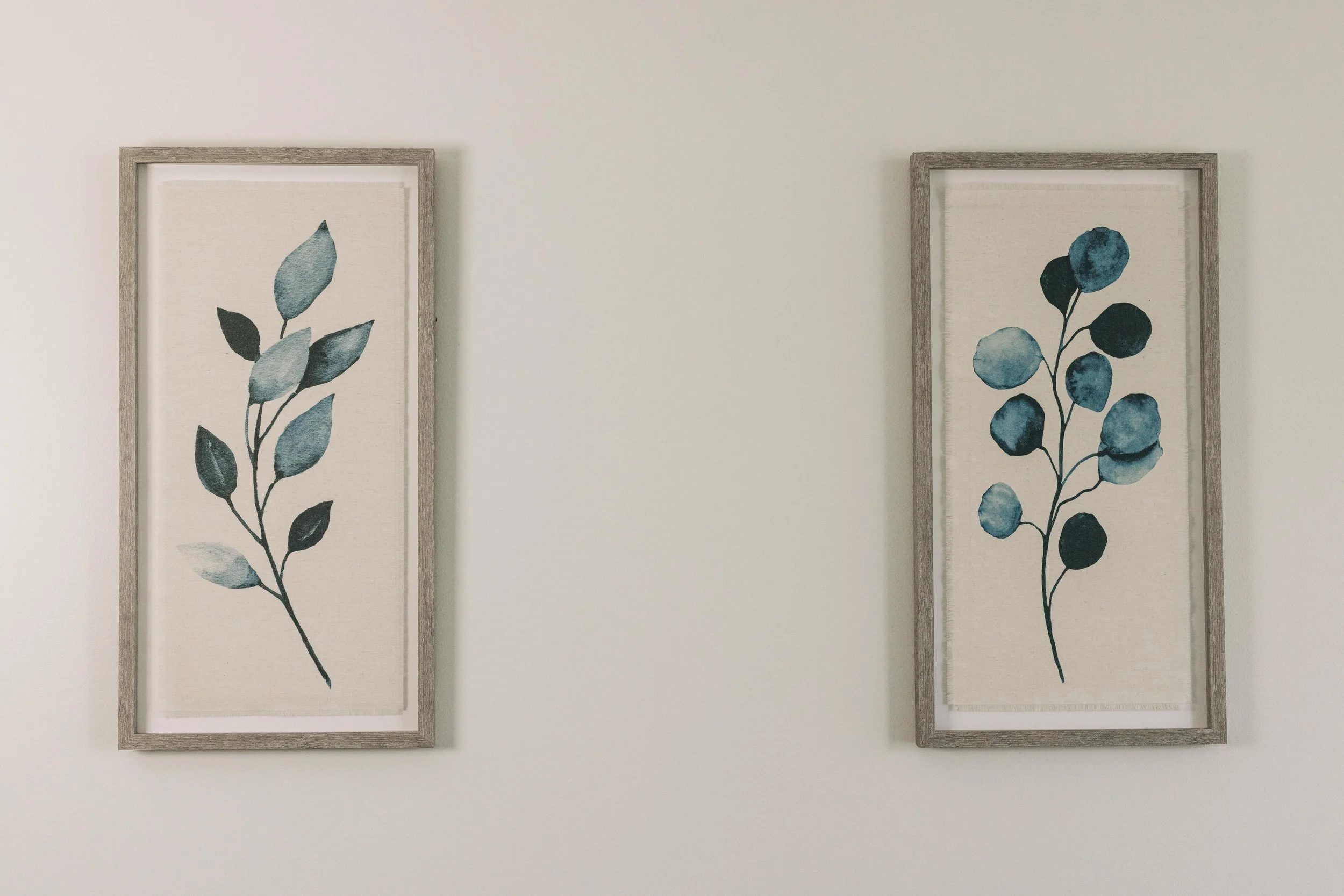 Two framed botanical prints featuring watercolor illustrations of blue and teal leaves, hung on an off-white wall.