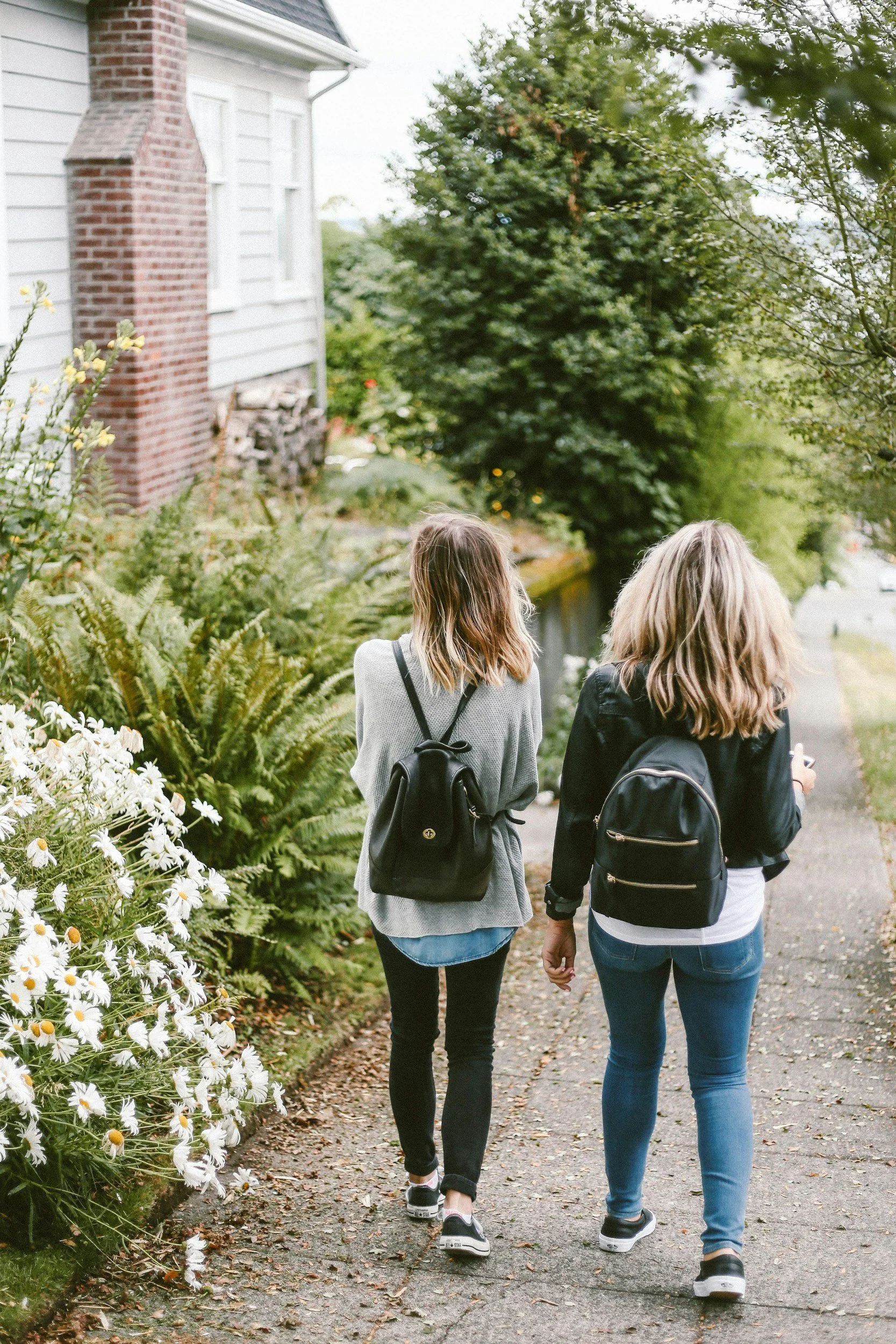 Two women with backpacks walking down a paved sidewalk surrounded by greenery and flowers, with a house and trees in the background.