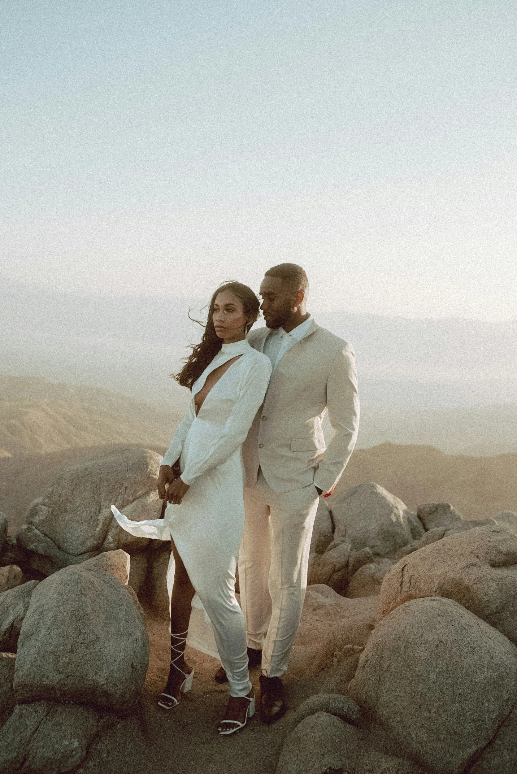 A man and a woman dressed in elegant white clothing standing among large rocks in a desert landscape at sunset.