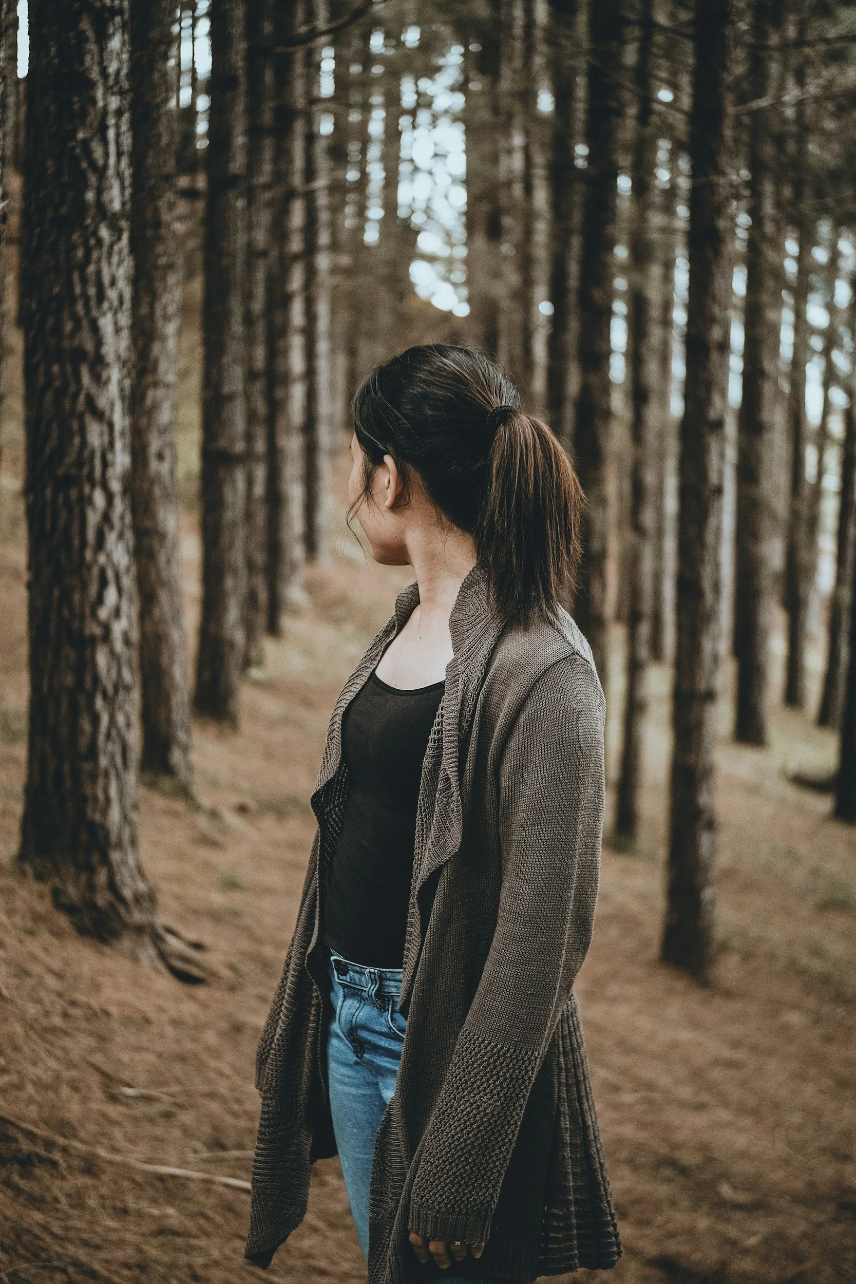 Woman standing in a forest with tall trees, wearing a dark top, a brown sweater, and jeans, with her hair tied back.