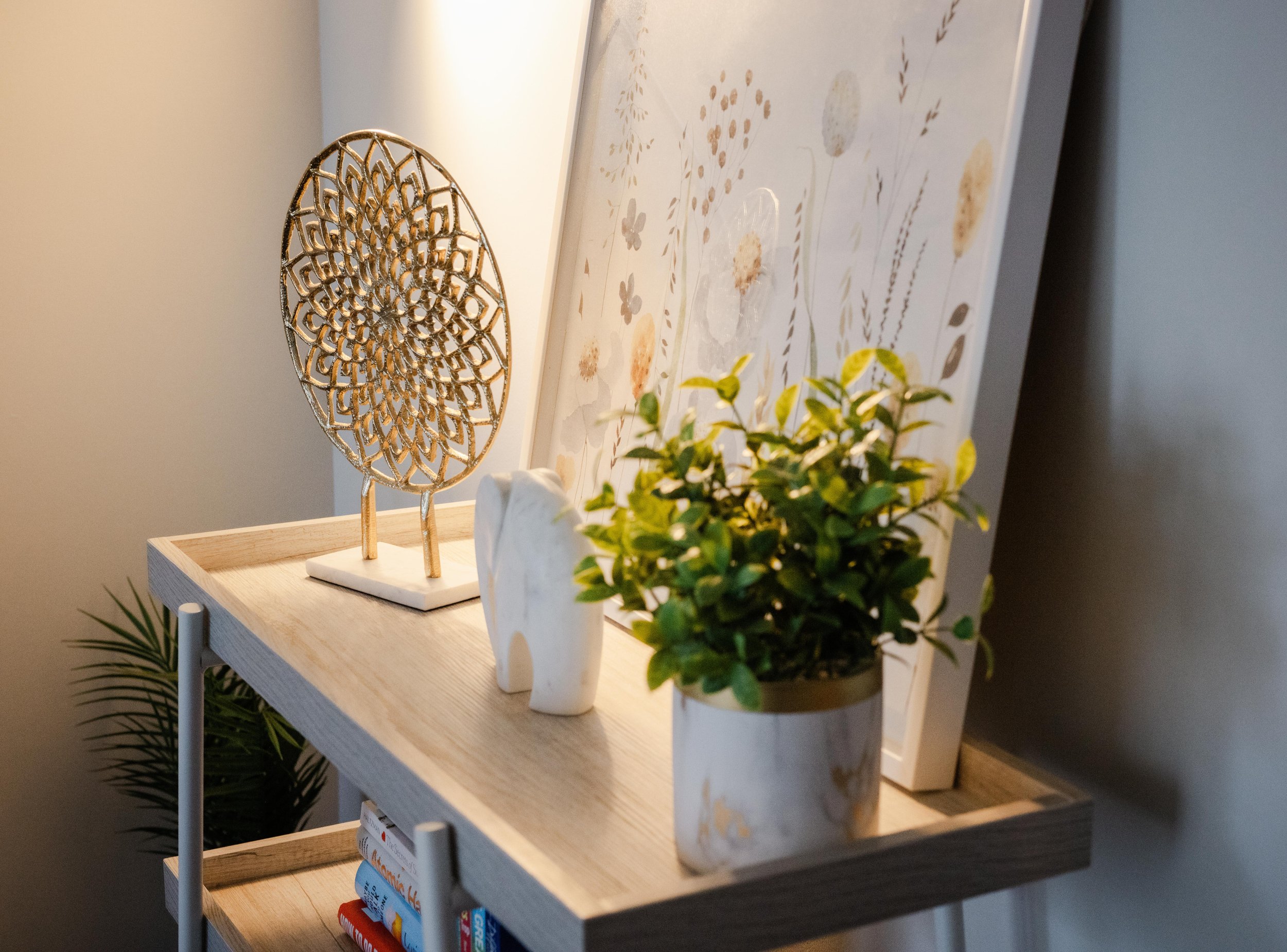A decorative metal sculpture on a wooden shelf, next to a potted plant and a ceramic animal figurine in a cozy, well-lit space with a floral wallpaper background.