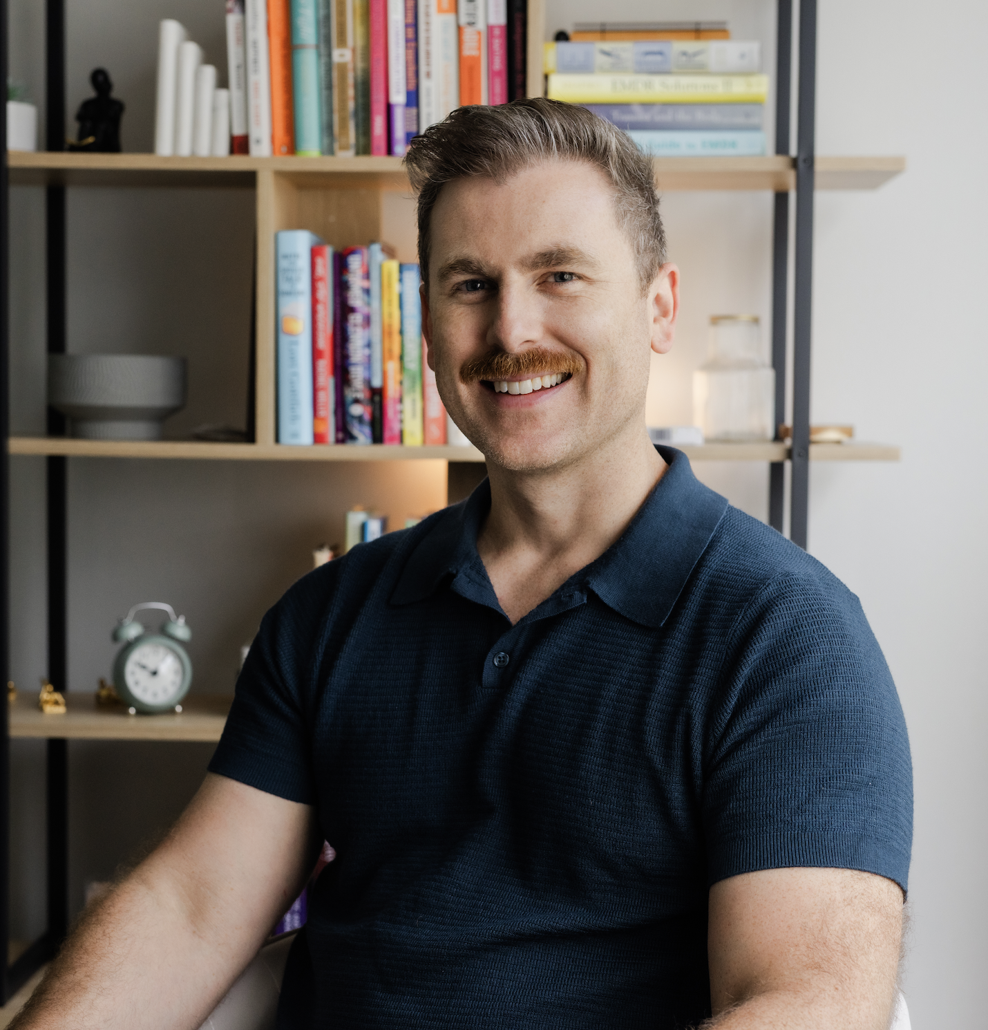 A smiling man with a mustache wearing a navy blue polo shirt, sitting in a room with a bookshelf filled with colorful books, a small clock, and decorative items.