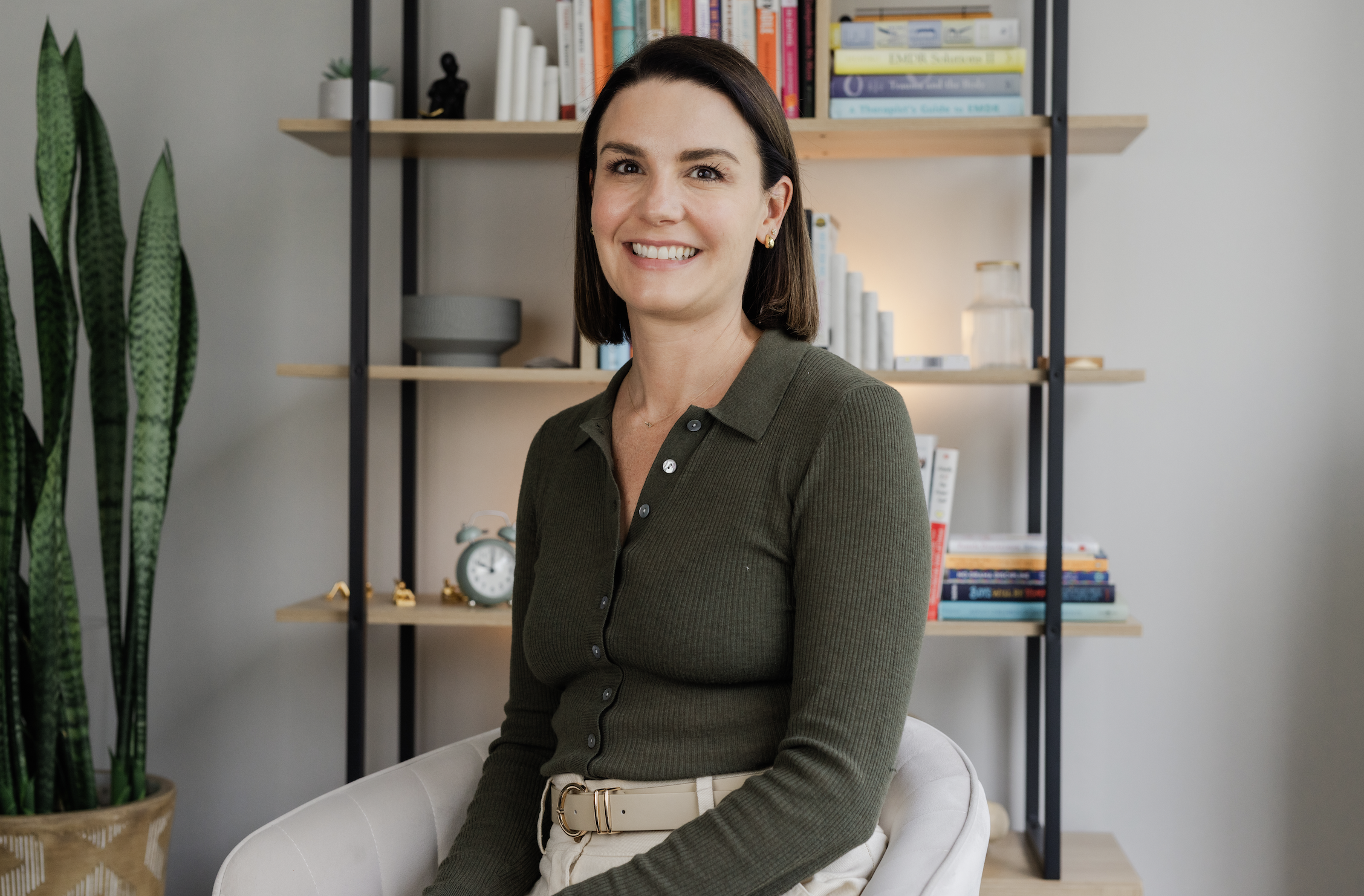 A woman with dark brown hair and light skin sitting on a white chair in front of a bookshelf, smiling. She is wearing a green button-up top and beige pants with a white belt.