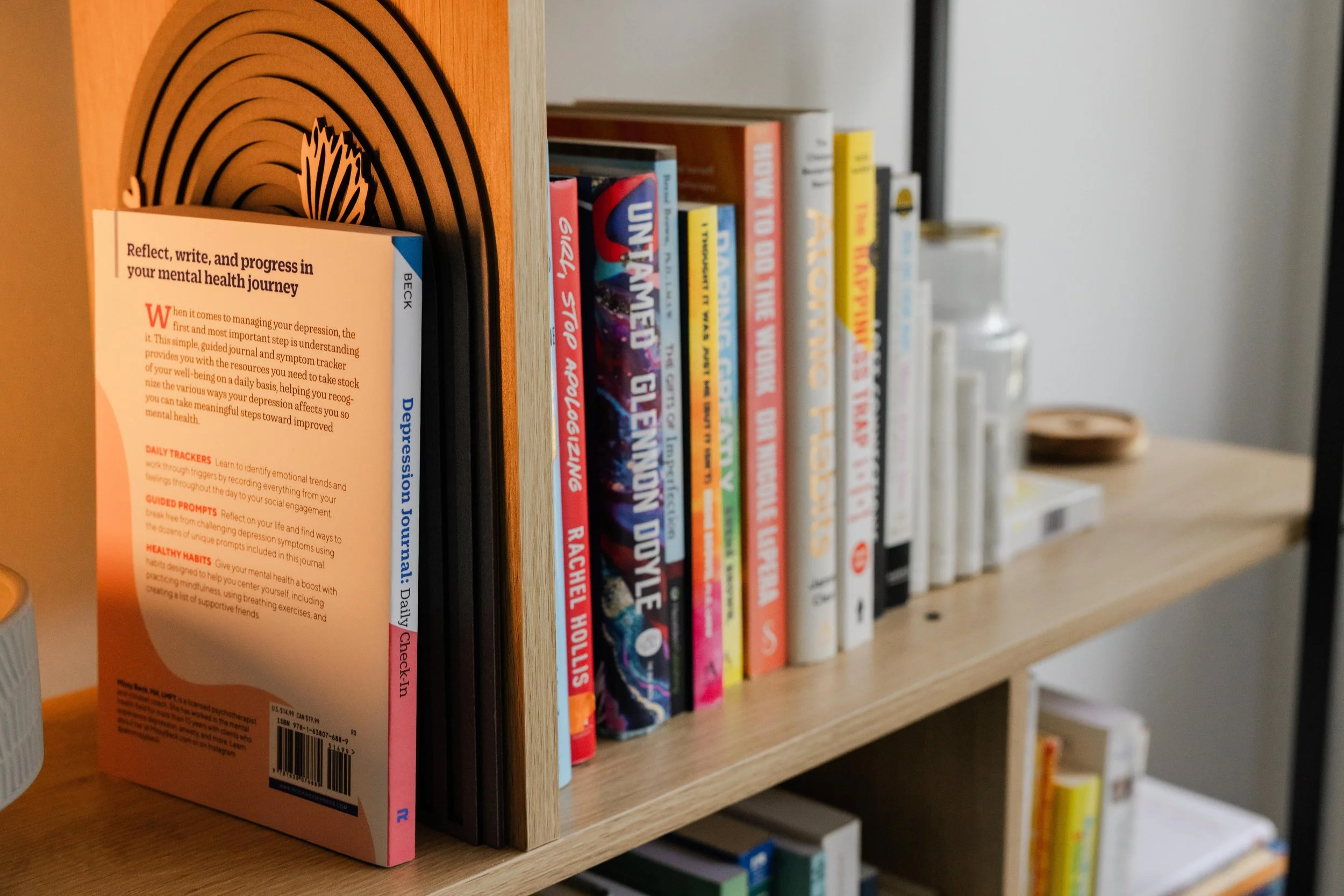 A bookshelf with a row of books, focusing on a specific book with an orange and pink cover. The books are standing upright on a wooden shelf, and the background shows other shelves with more books.
