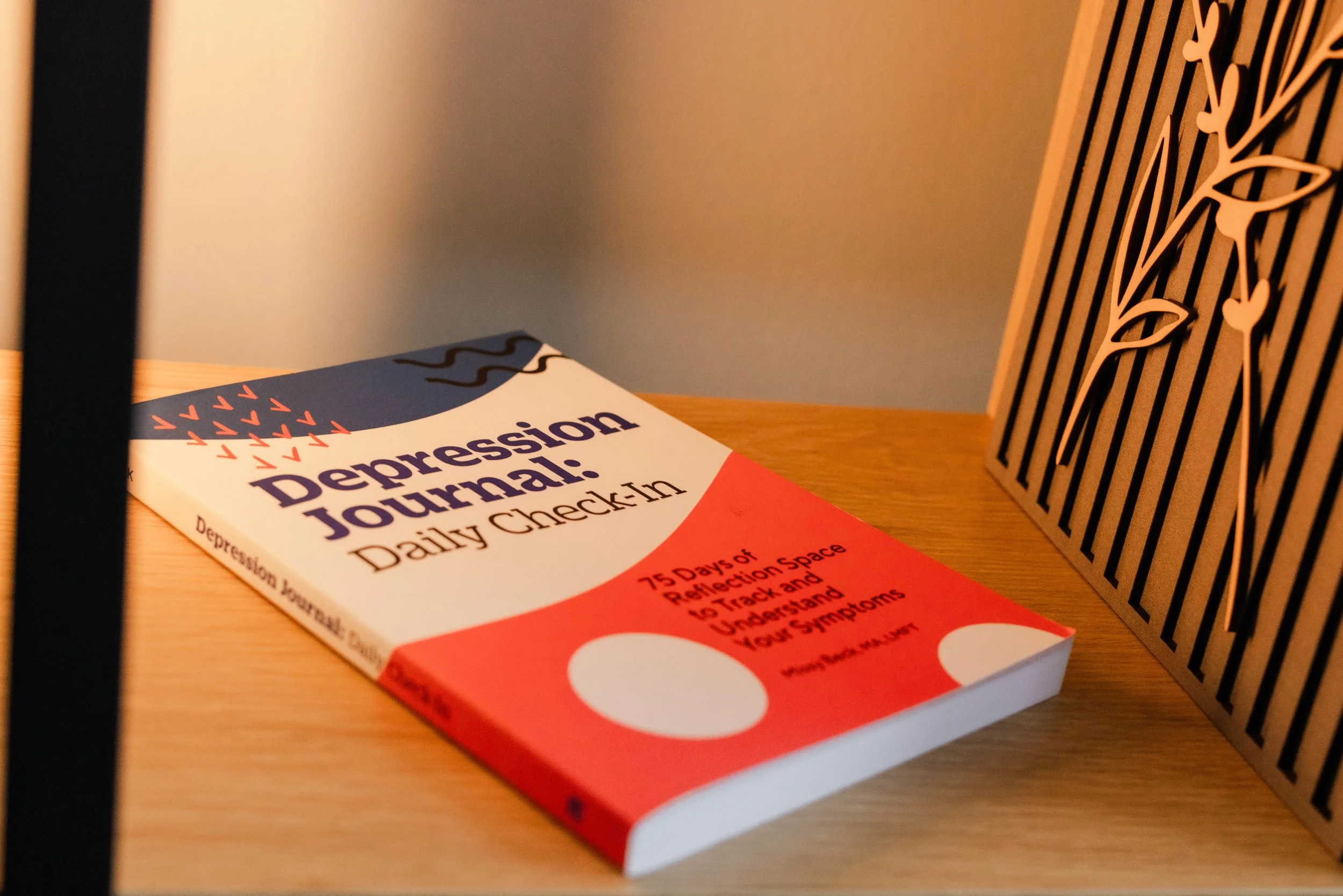 A book titled "Depression Journal: Daily Checklist" placed on a wooden surface next to a decorative black metal panel with a leaf pattern, with a gray wall in the background.