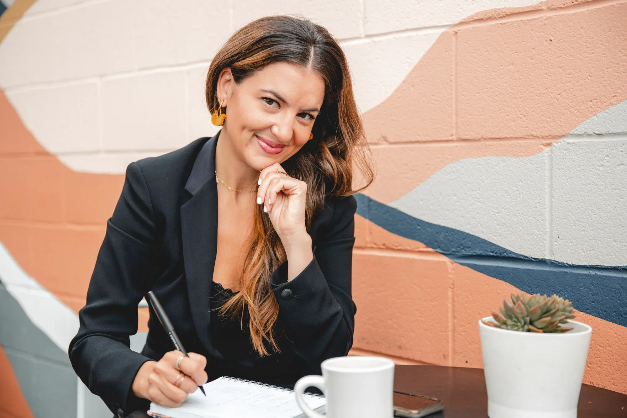 A woman with long brown hair, wearing a black blazer and gold earrings, is sitting at a table with coffee mugs and a potted succulent plant. She is smiling, looking at the camera, and writing in a notebook with a pen. The background features a colorful mural painted on a brick wall.