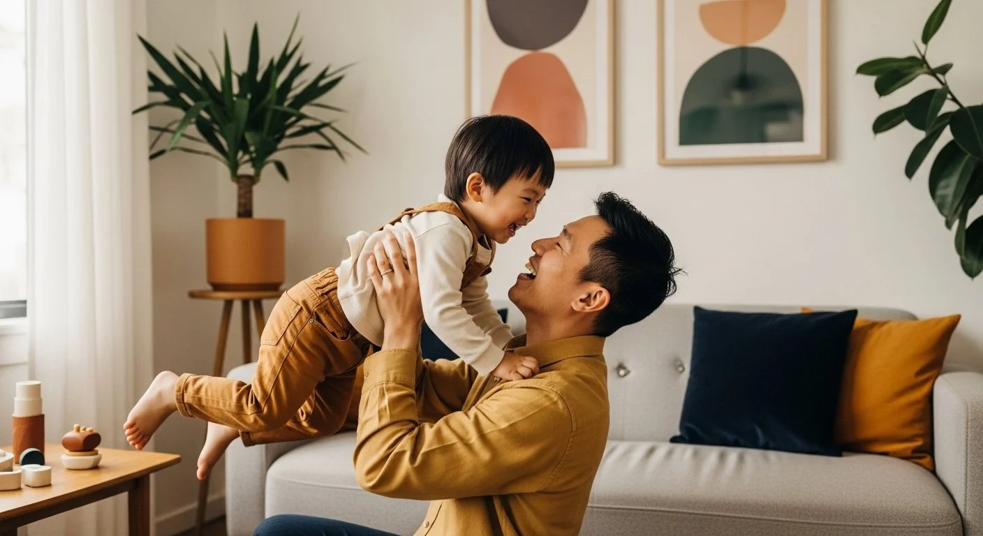 A man and a young boy are playing and laughing together in a living room, with the man lifting the boy into the air. There are plants, framed artwork, and cushions in the background.