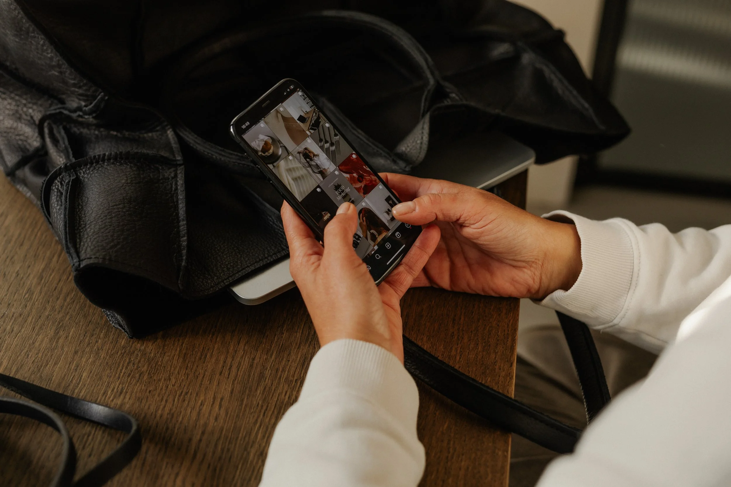 A person holding a smartphone and browsing through photos on Instagram, placed on a wooden table next to a black leather bag.