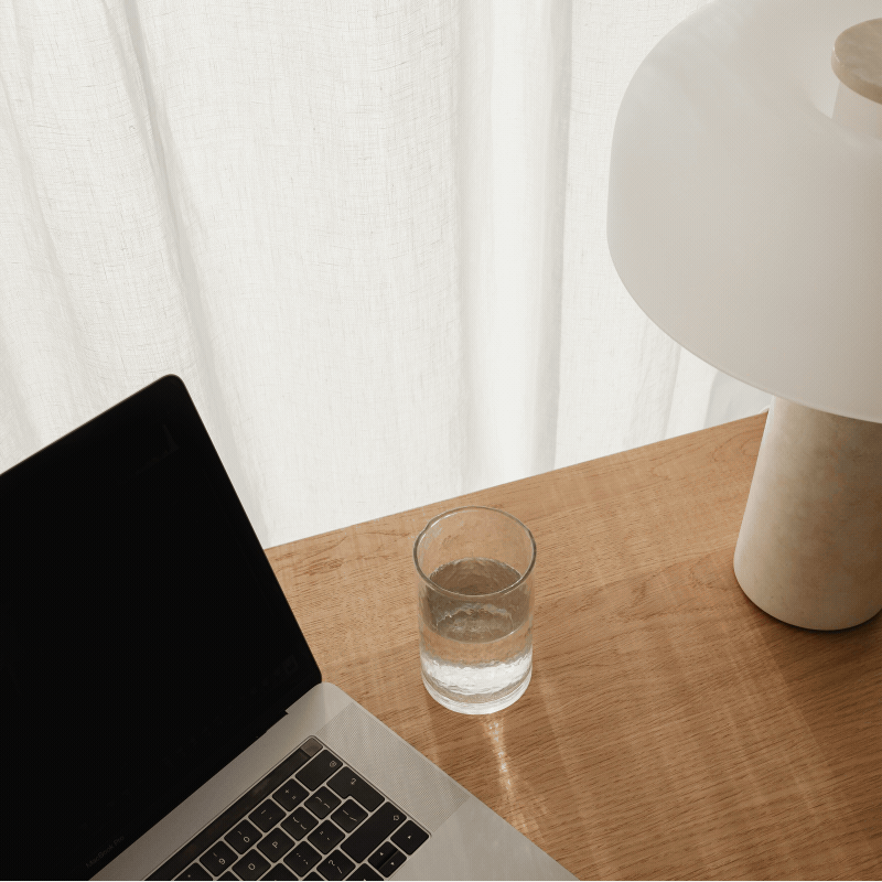 A close-up of a wooden desk with a laptop, a glass of water, and a white table lamp next to a window with sheer curtains.