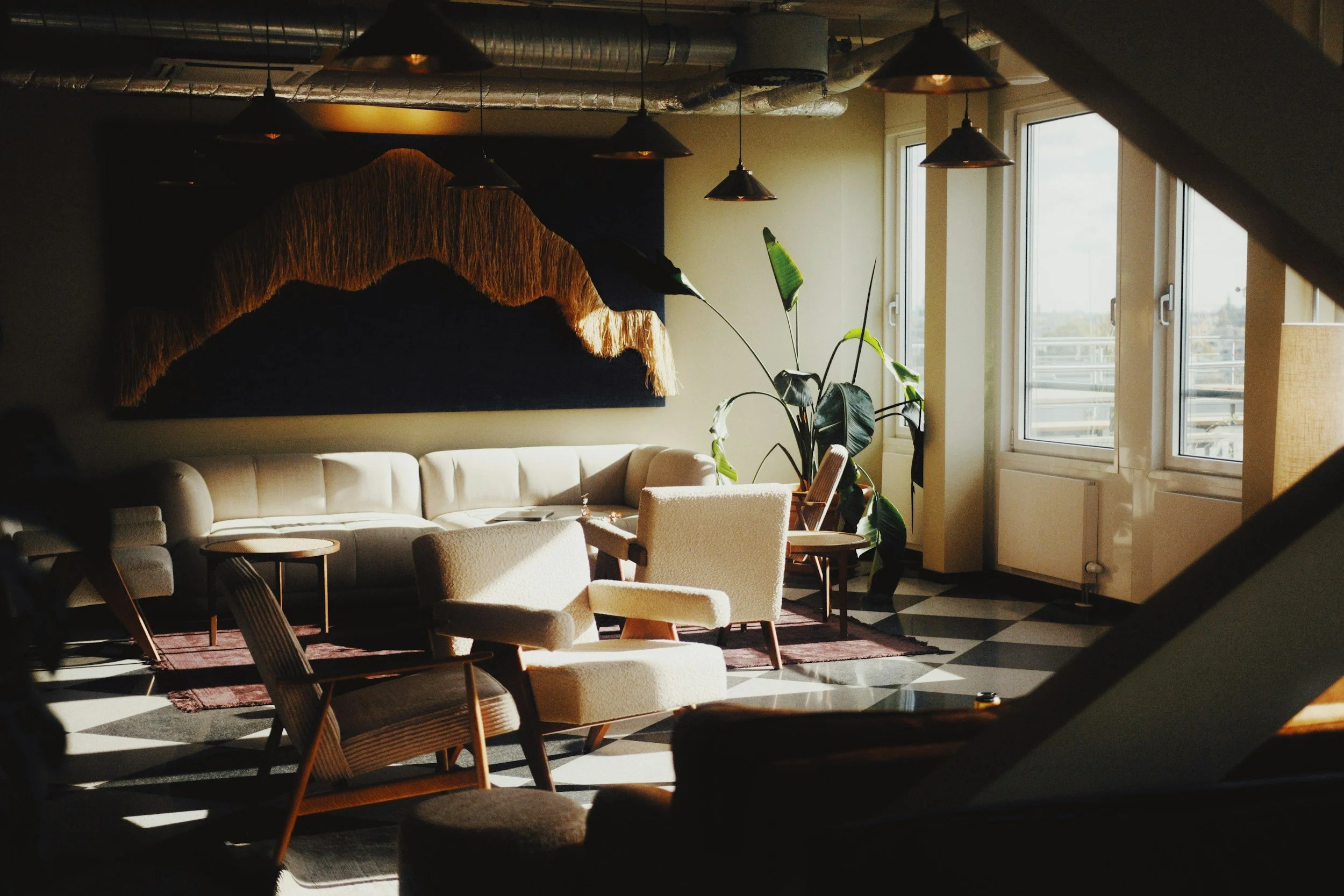 Sunlit modern lounge area with beige sofa, armchairs, large green plant near windows, geometric checkered floor, and hanging pendant lights, featuring a wall art piece.