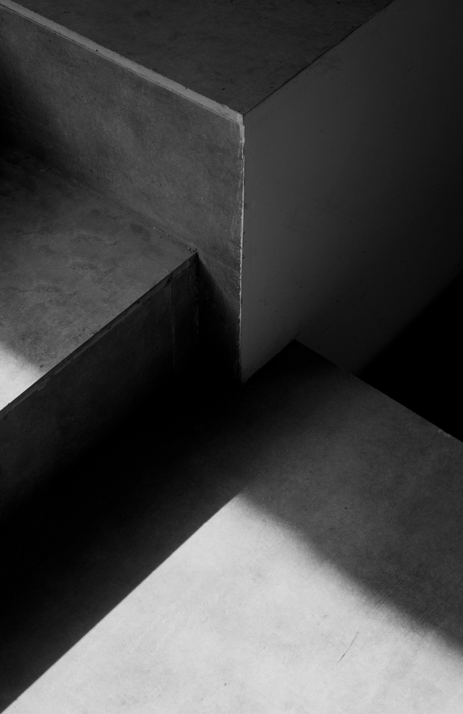 A black and white photograph of a geometric corner where a wall, floor, and ceiling meet, with shadows casting across the surfaces.