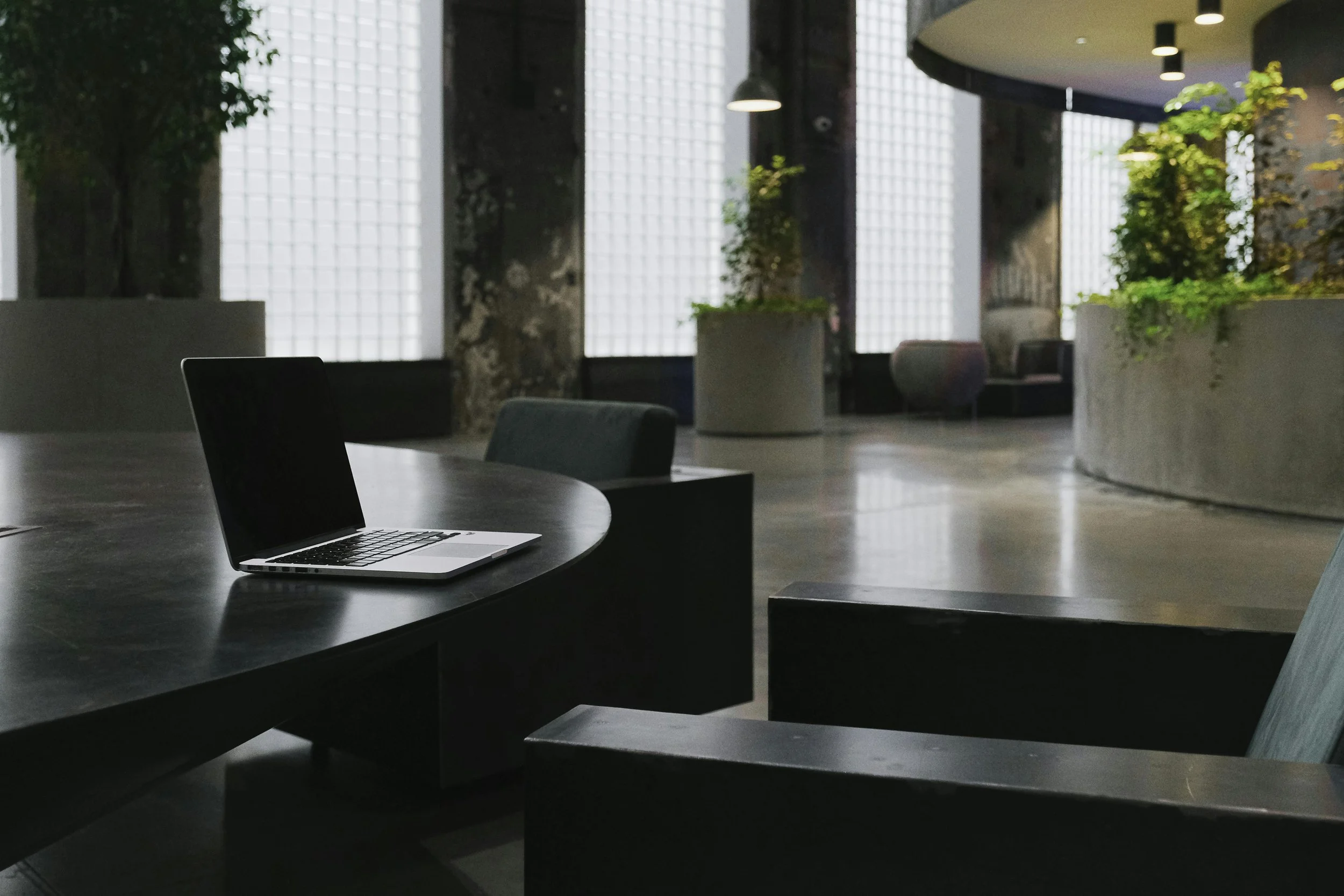 A modern, quiet office or lounge area with a black table, a laptop, and dark green chairs. Large windows with frosted glass blocks allow natural light, and there are tall planters with green plants. The space has concrete floors, exposed columns, and a curved wall with additional seating.