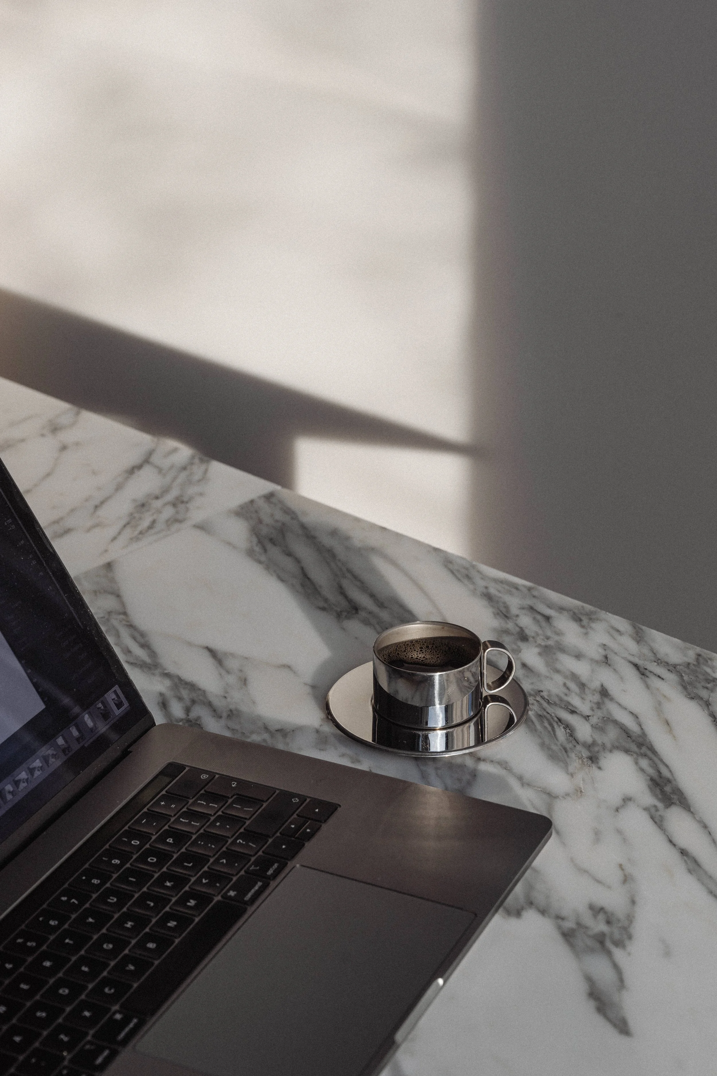 Laptop on a marble table with a cup of coffee on a saucer nearby.