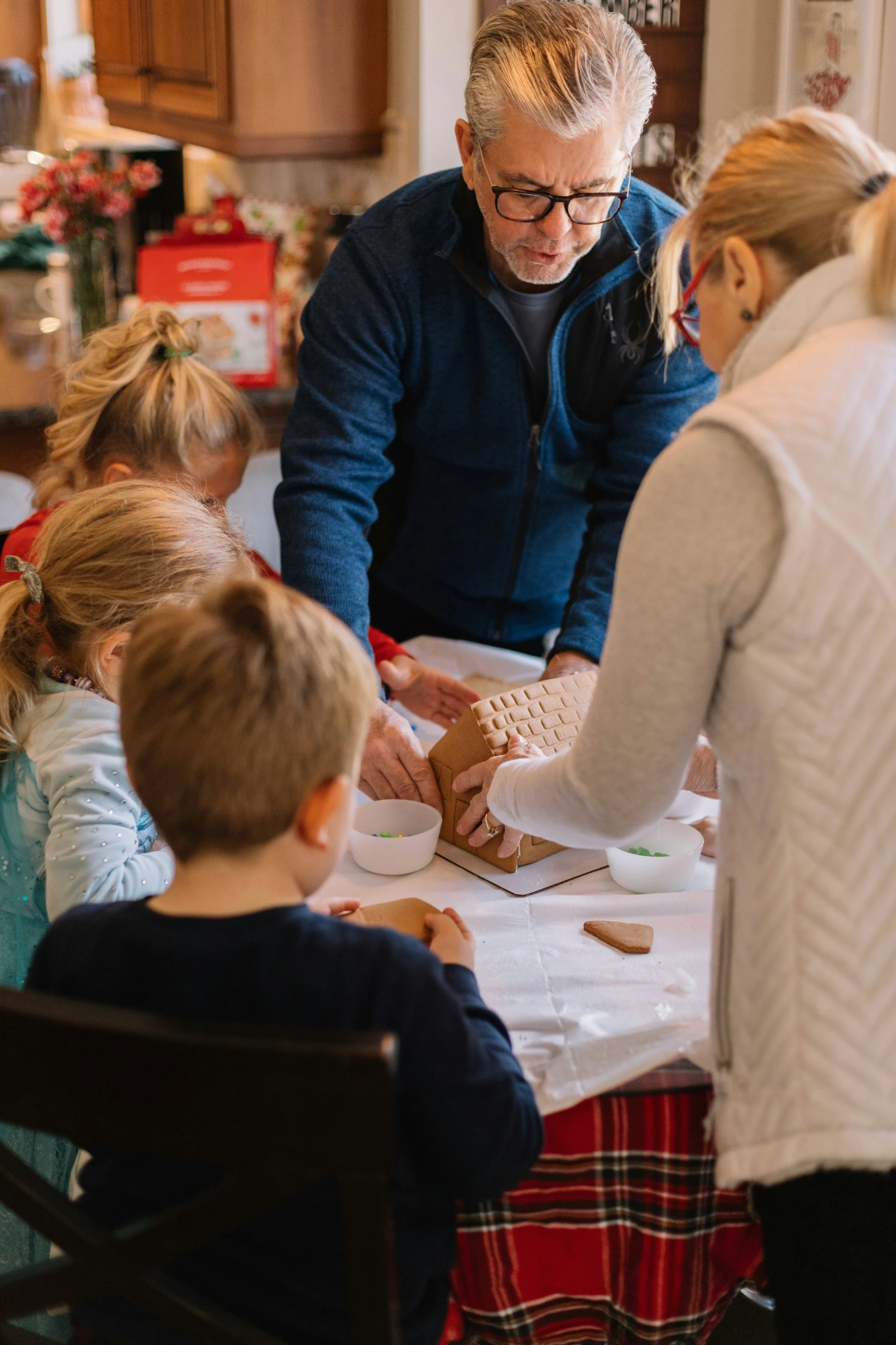 Family gathered around a table decorated for Christmas, decorating gingerbread cookies with gingerbread house in the background.