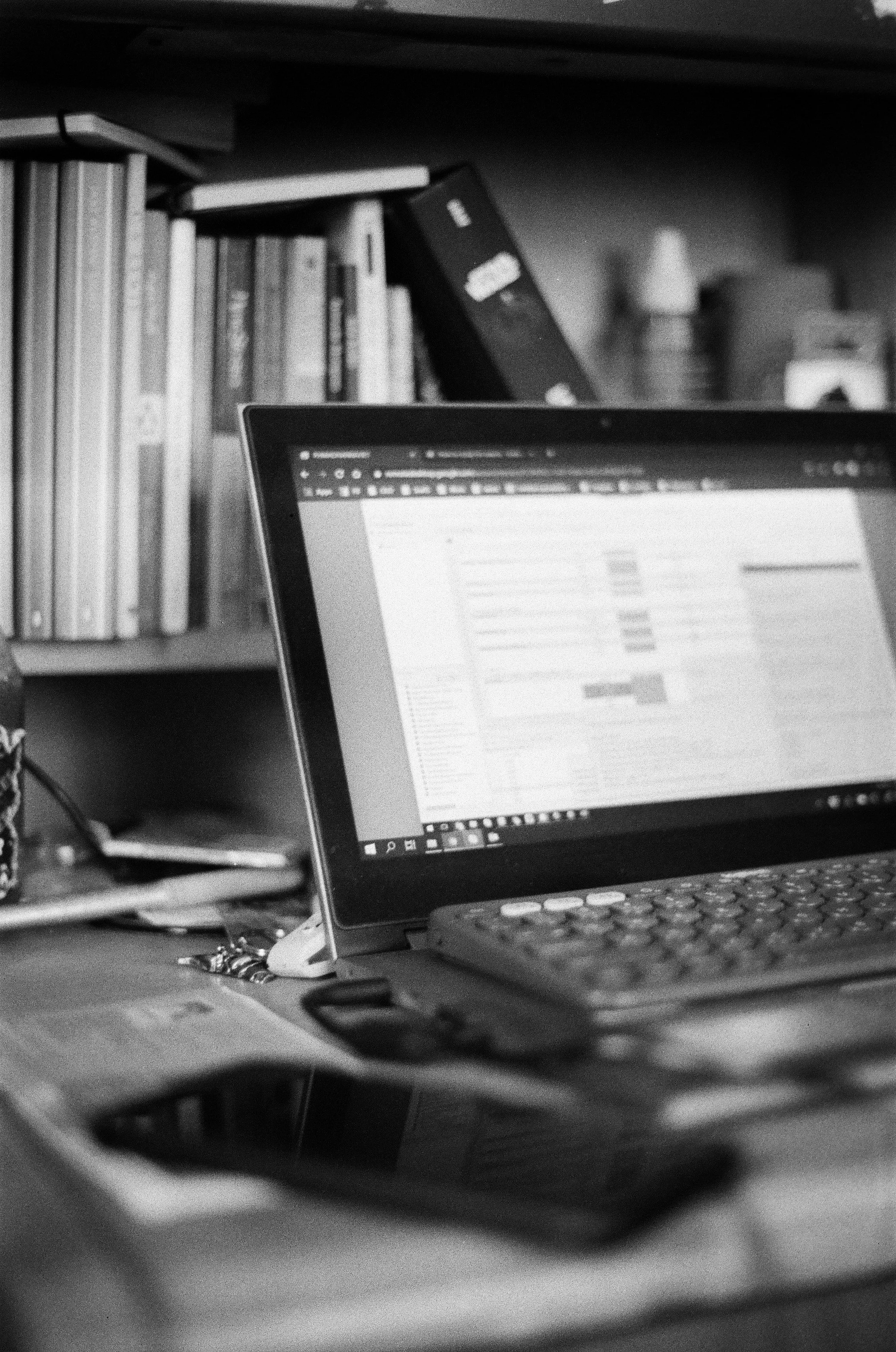 A black and white photo of a cluttered desk with a laptop, smartphone, and books in the background.