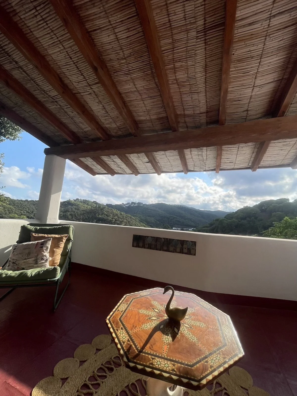 Outside balcony with mountain view, shaded by a wooden and bamboo roof, featuring a green cushioned lounge chair with pillows, a decorative table with a metal duck sculpture, a beige mat, and a small stained glass window on the wall.