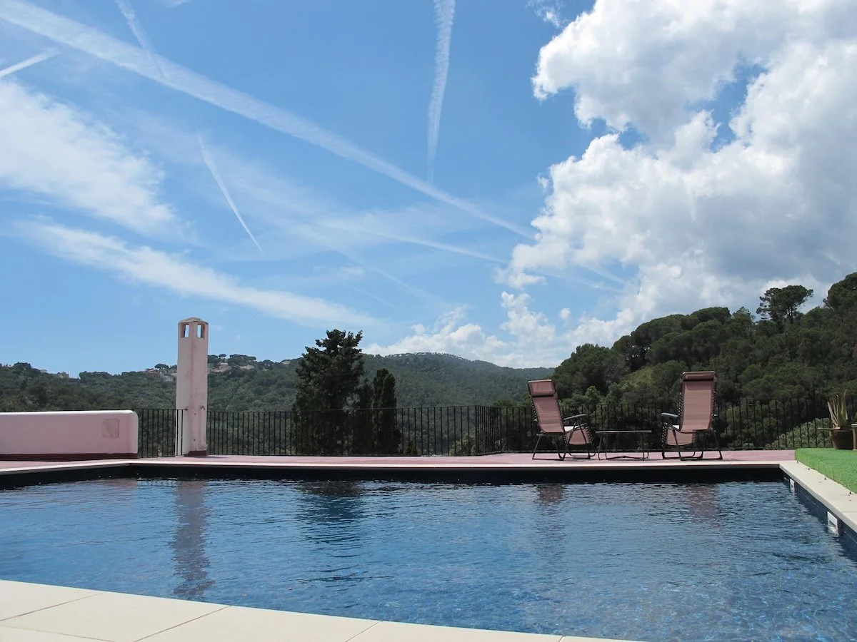 A backyard swimming pool with lounge chairs on a patio, overlooking green hills and trees under a blue sky with scattered clouds and contrails.
