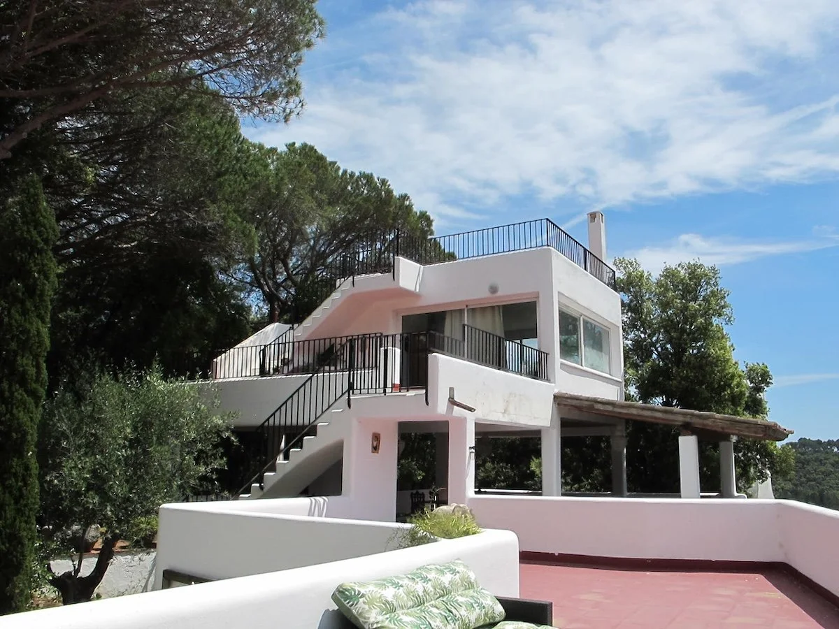 A white modern house with multiple levels and outdoor staircases, surrounded by trees, under a partly cloudy blue sky.