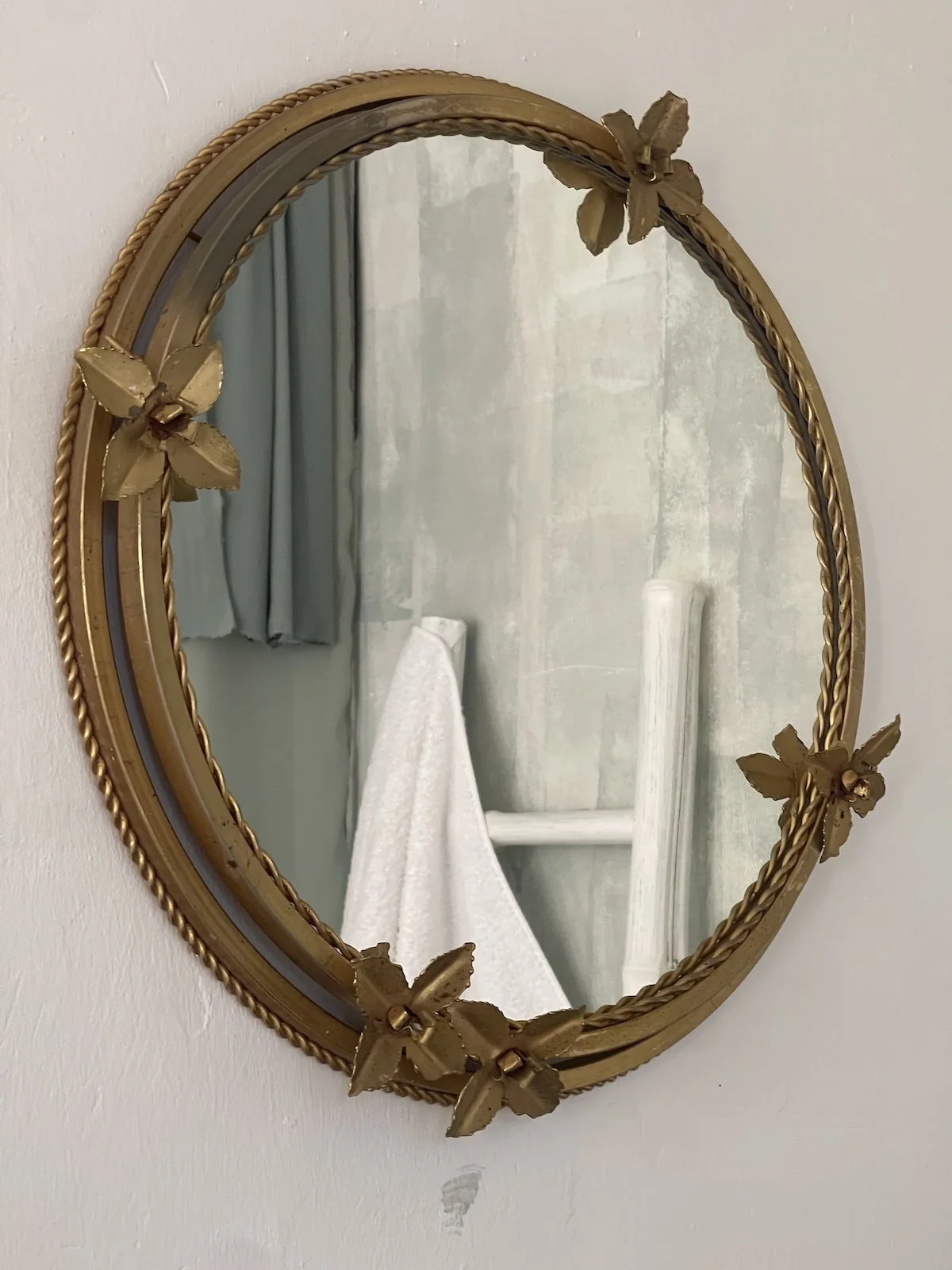 A decorative oval mirror with a gold floral frame hanging on a white wall, reflecting a bathroom with a white towel on a rack and a pale green curtain.