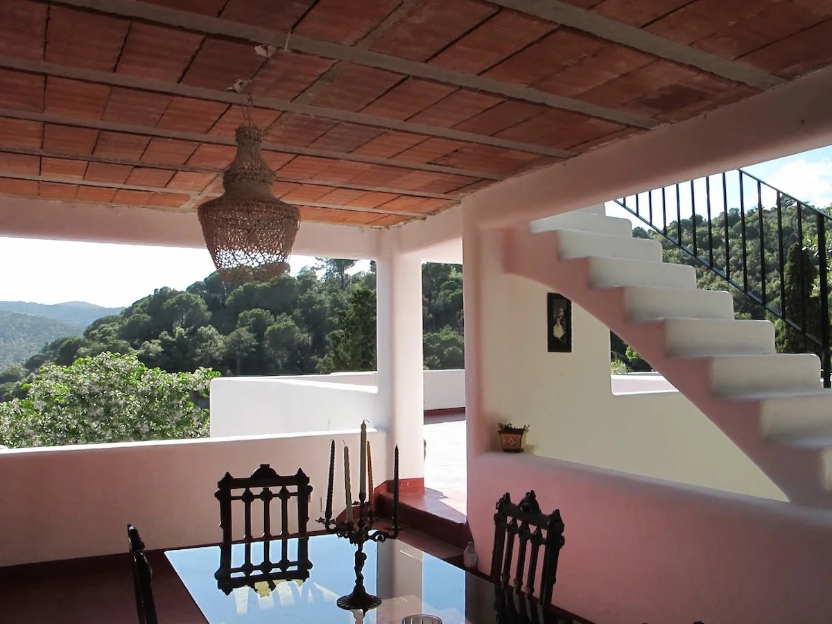 Interior view of a room with a dining table, chairs, hanging chandelier, staircase, potted plant, and scenic mountain view outside.