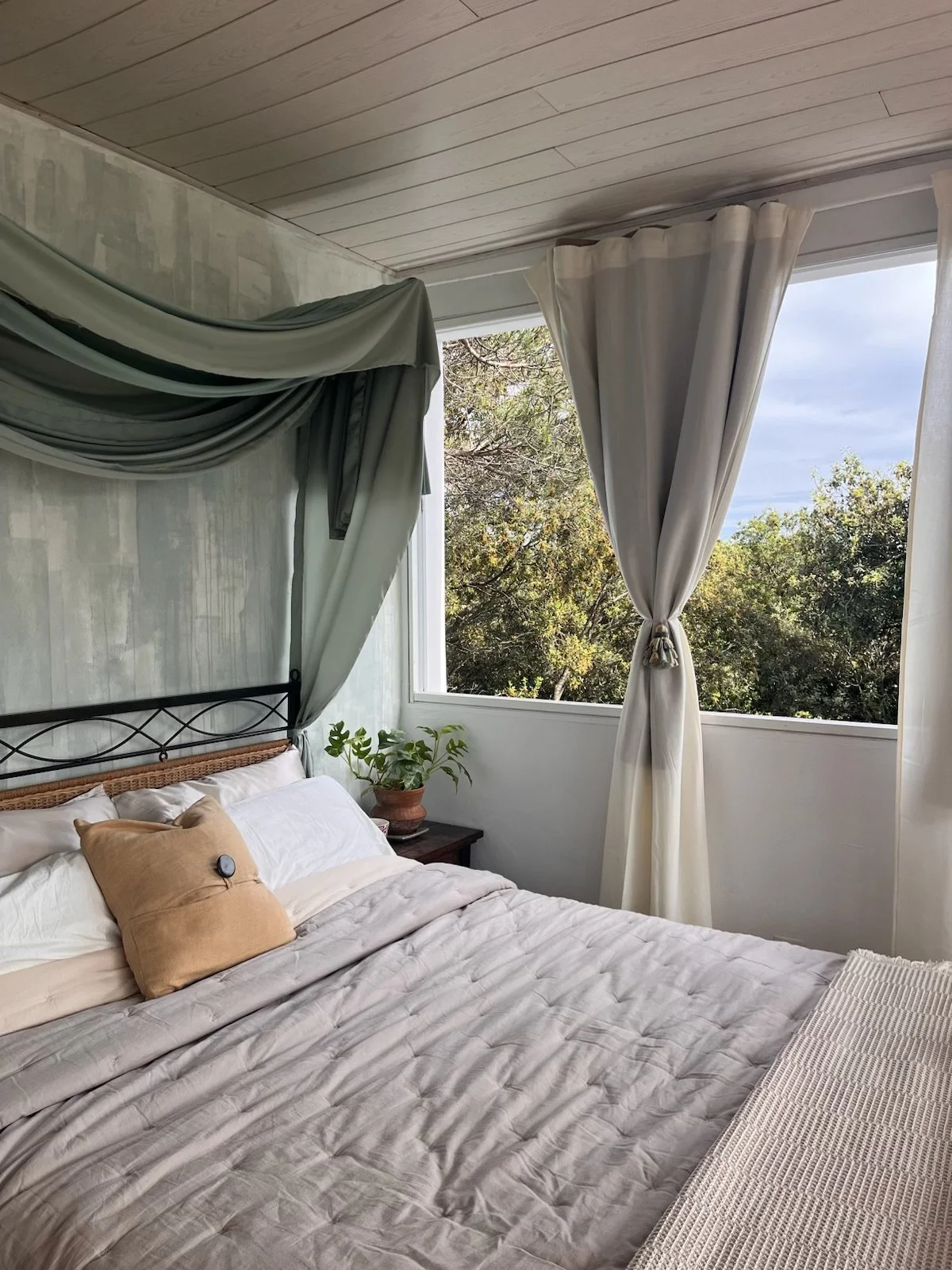 Bedroom with bed, pillows, beige quilt, a black metal headboard, a side table with a potted plant, large window with white curtains tied back, and trees outside.
