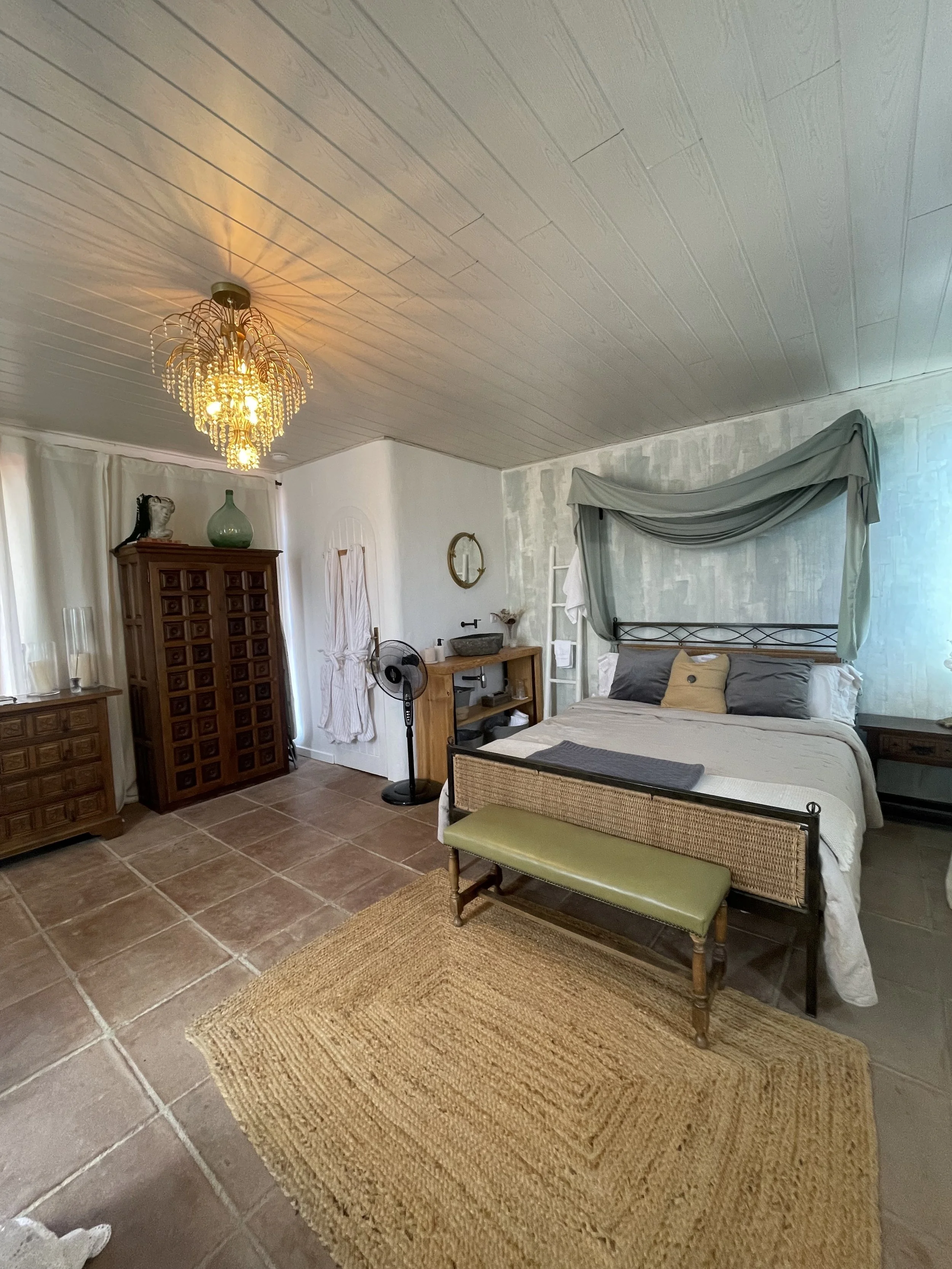 A bedroom with a bed, a green bench at the foot of the bed, a wooden cabinet, a chandelier, and a terracotta tile floor with a woven rug. There is a fan, a mirror, and various decorative items in the room.