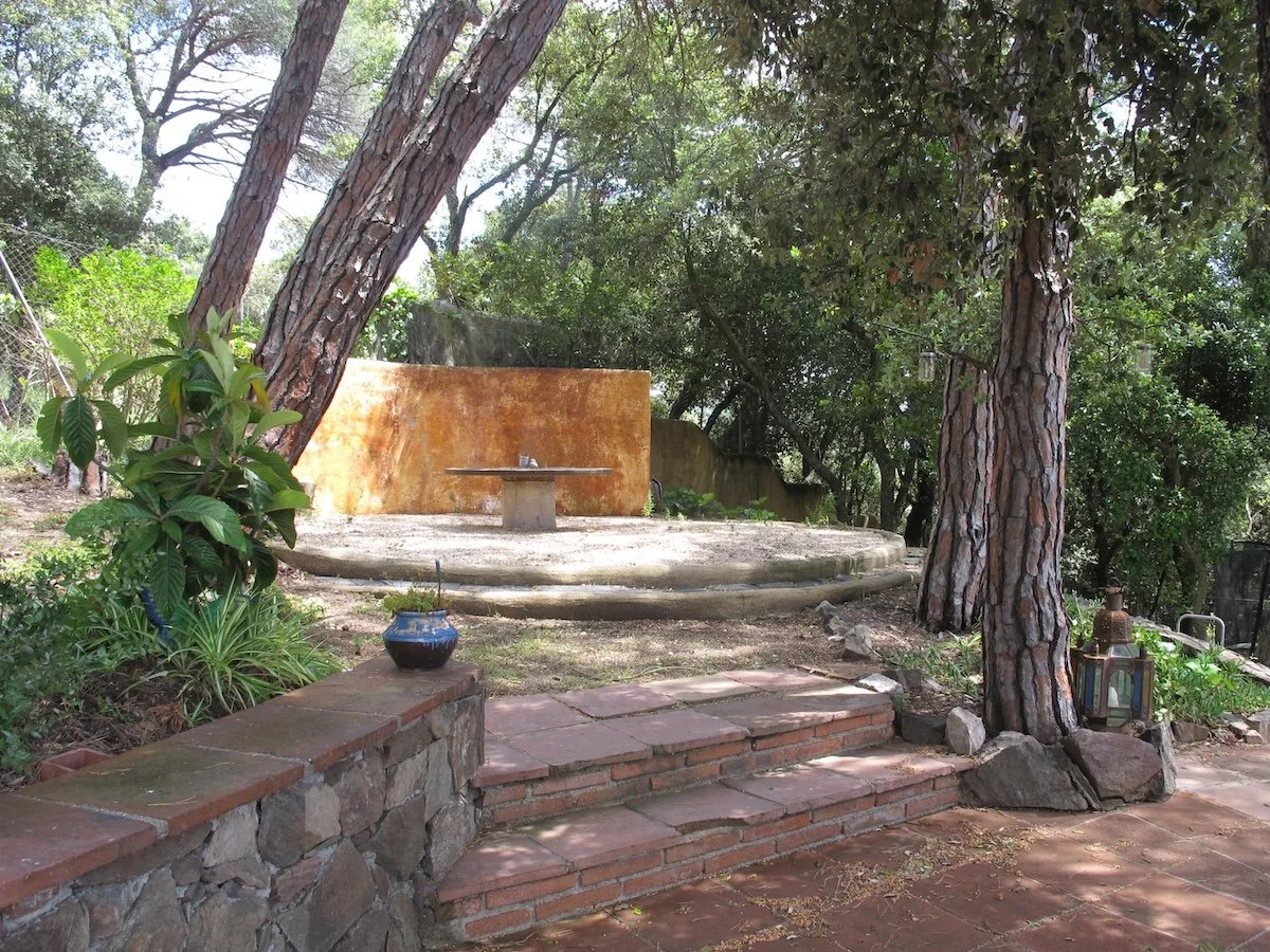 Outdoor scene with steps leading up to a cement platform, surrounded by trees and plants, with a rusty wall in the background and a small blue pot on the brick wall in the foreground.