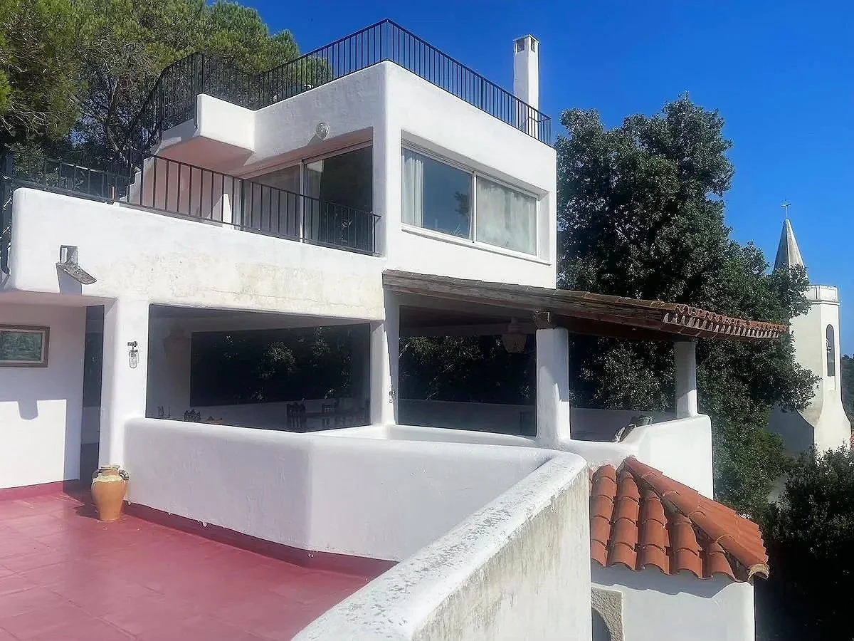 White multi-level house with terracotta roof tiles, large glass windows, black metal railings, and a pink tiled patio under a bright blue sky with trees in the background.