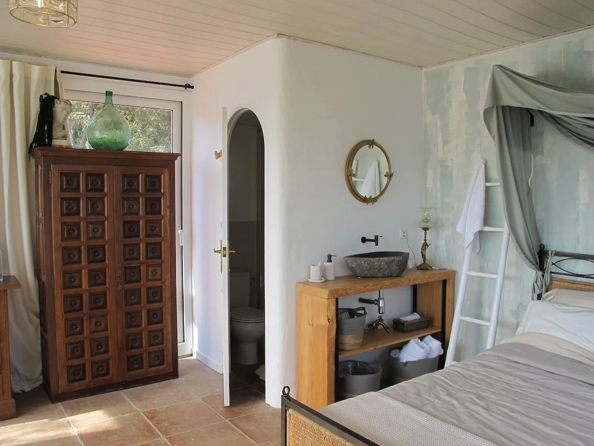 A cozy bedroom with a partial view of a bed, a wooden vanity with a stone sink, a mirror, a wooden cabinet, and a small bathroom with a toilet visible through an open door. There is a ladder with a towel hanging on it and a large glass bottle on top of the cabinet.
