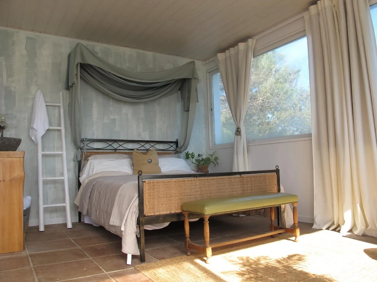 A cozy bedroom with a bed featuring a black metal headboard, a beige bedspread, and white pillows. There is a small green bench at the foot of the bed, a ladder shelf with a towel on it, and a potted plant on the nightstand. Large windows with white curtains allow natural light to fill the room.