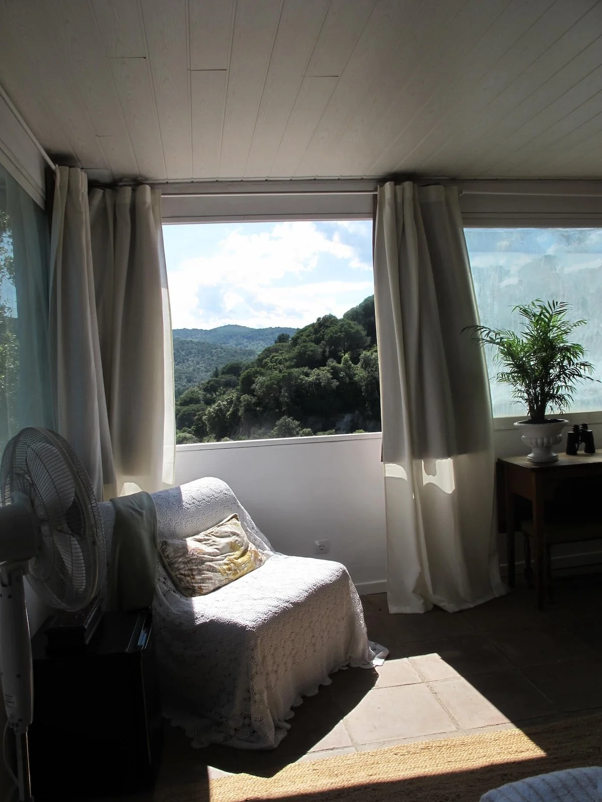 Living room with open window showing green hills and a partly cloudy sky, beige curtains, a white covered couch with a pillow, a table with a potted plant, and an electric fan.
