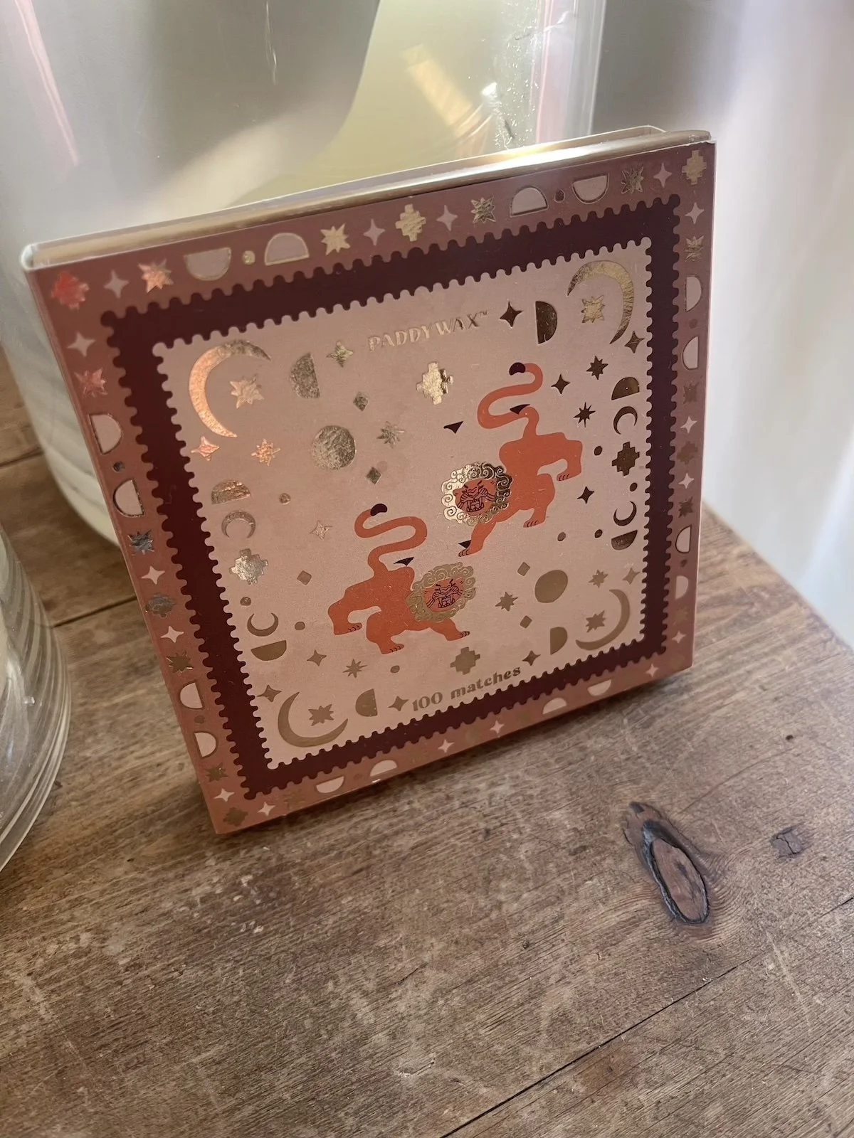 A decorative box with pink and gold moon, star, and lion illustrations, labeled 'Paddy Wax' and '100 matches' on a wooden surface.