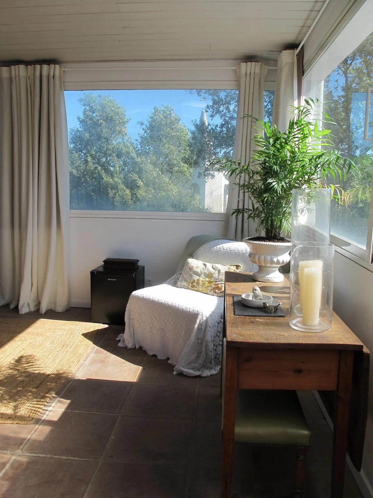 Sunlit interior room with large window showing trees. A sofa with a white lace cover and a decorative pillow, a wooden table with a large potted plant, candles, and decorative items. Curtains frame the window, and a small black cabinet is nearby.