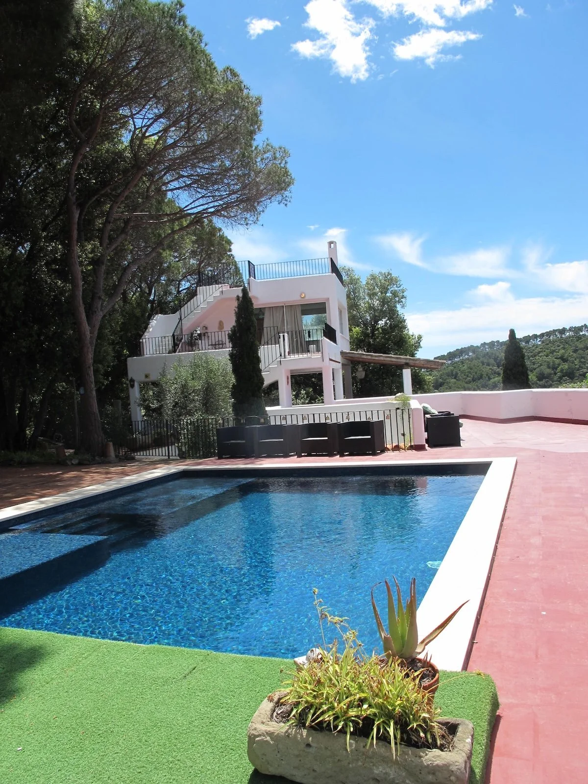 A luxurious backyard with a swimming pool, an outdoor seating area, and a white, multi-level house with balconies and stairs, surrounded by trees and under a bright blue sky with scattered clouds.