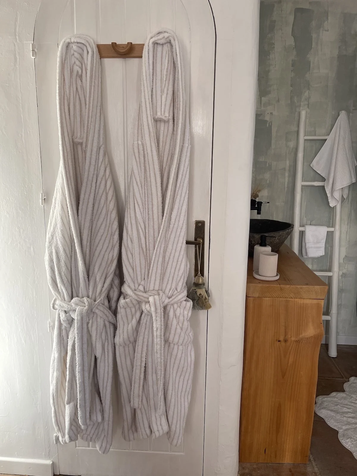 Two plush, white robes hanging on a wooden hook on a white door, with one facing forward and the other facing backward, tied at the waist. To the right, there is a bathroom vanity with a dark stone sink, a soap dispenser, and a white decorative item. A white towel is hanging on a ladder-style towel rack on the wall behind the vanity.