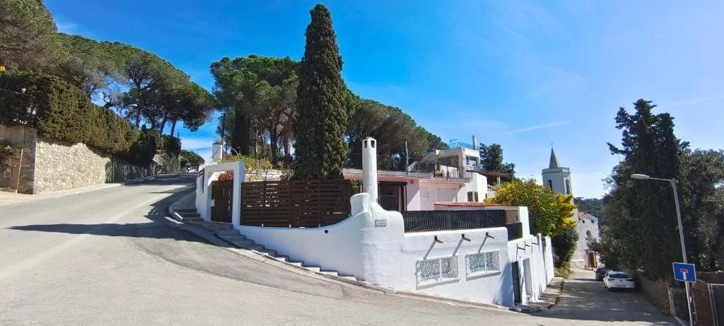 A residential street with a house on a hillside, white walls, and a tall chimney, with trees and a clear blue sky in the background.