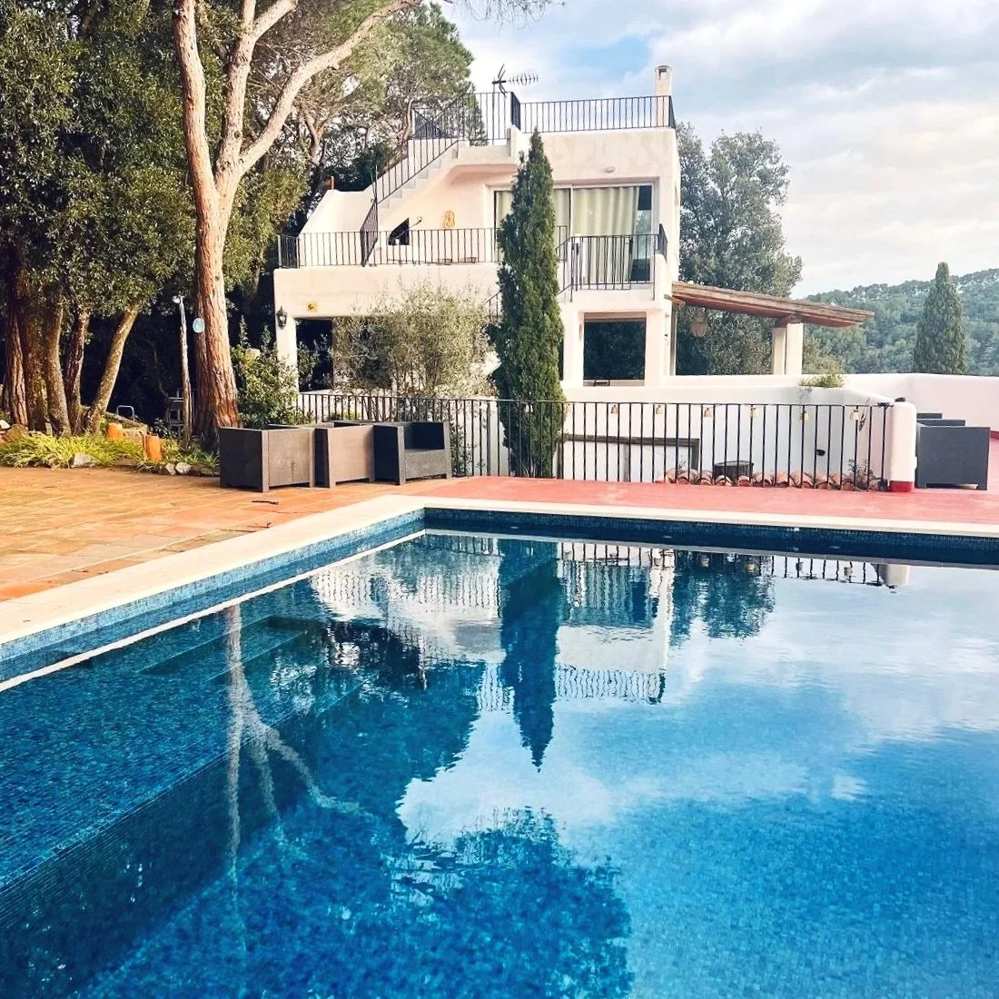 A swimming pool outdoors with a white house with balconies in the background. There are trees and outdoor furniture around the pool area.