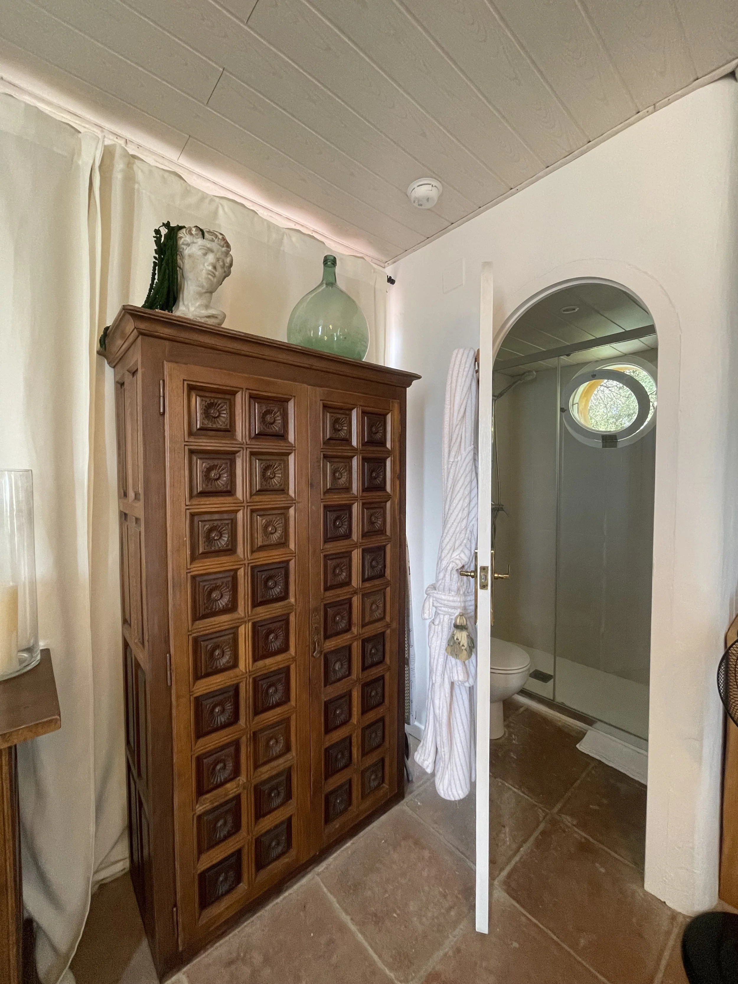 A wooden cabinet with carved square patterns on the doors is positioned near a shower area with glass doors. On top of the cabinet, there is a bust sculpture with some green plant or hair accessory draped over it and a large round glass bottle. To the right, a partial view of a bathroom with a toilet, shower, and a porthole window is visible, with a white curtain hanging next to the cabinet.