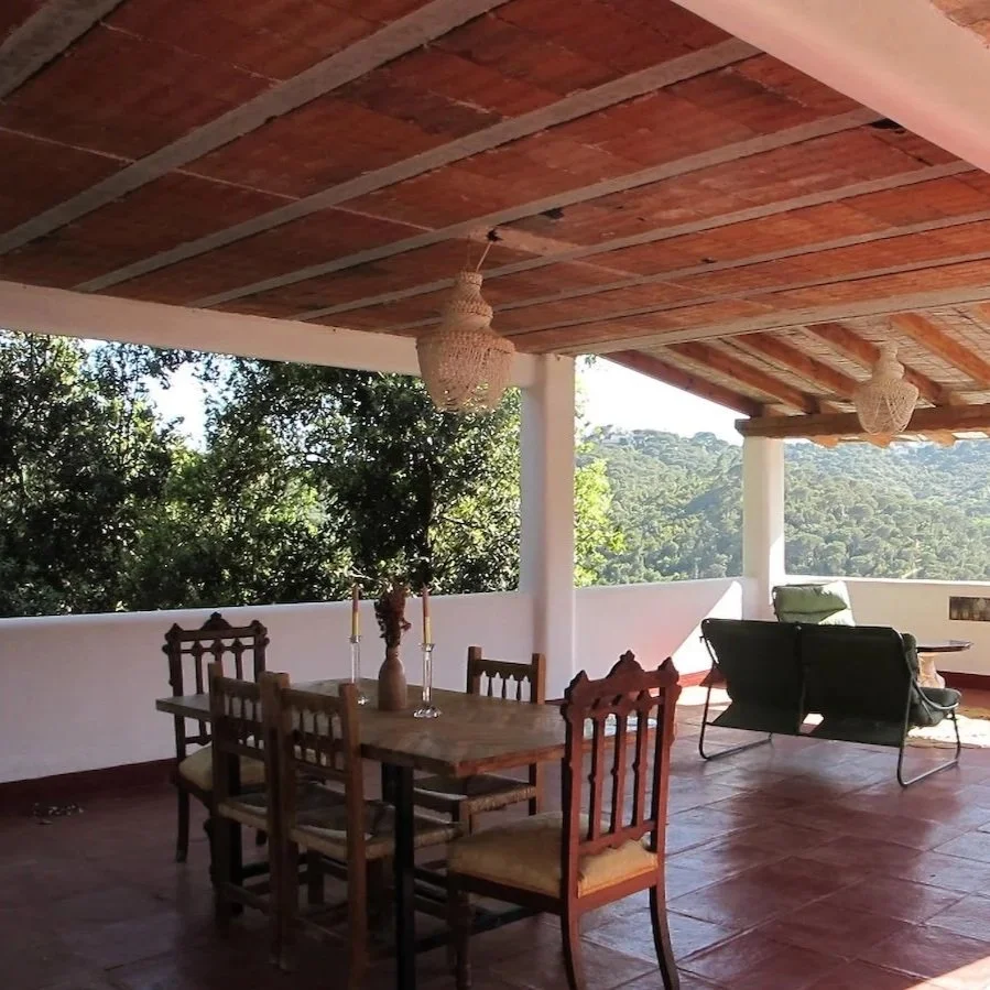 Covered outdoor patio with wooden ceiling, wooden dining table with six chairs, two candles, and a vase with flowers; two hanging wicker light fixtures; some lounge chairs and a scenic view of trees and hills in the background.