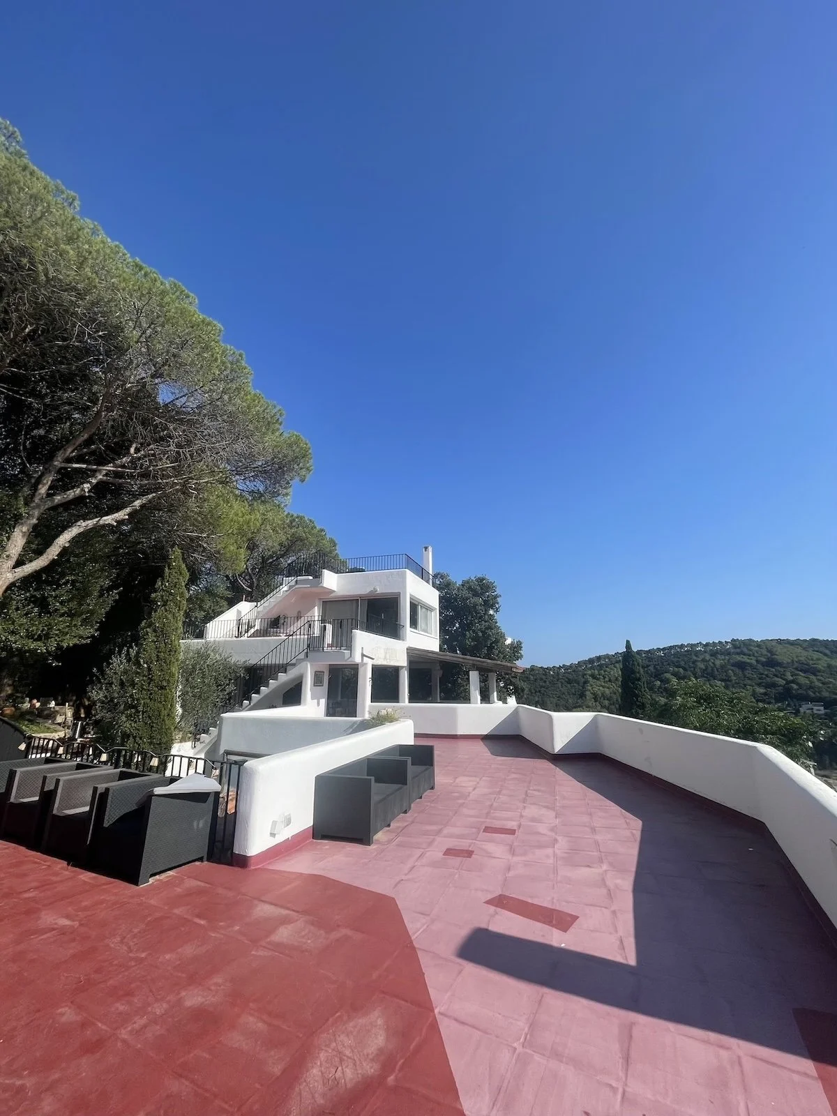 A spacious outdoor terrace with pink flooring, white walls, and black outdoor furniture, overlooking green hills under a clear blue sky.