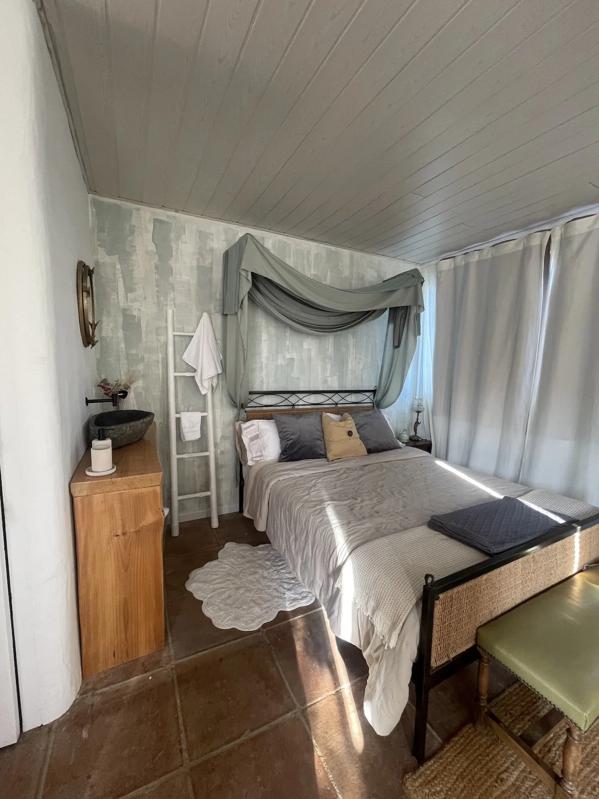 A cozy bedroom with a bed featuring gray and beige pillows and a beige bedspread, decorated with a canopy of draped gray fabric above the headboard, a small wooden side table with a lamp, a window with sheer curtains letting in sunlight, a white ladder with a towel, a small stone sink