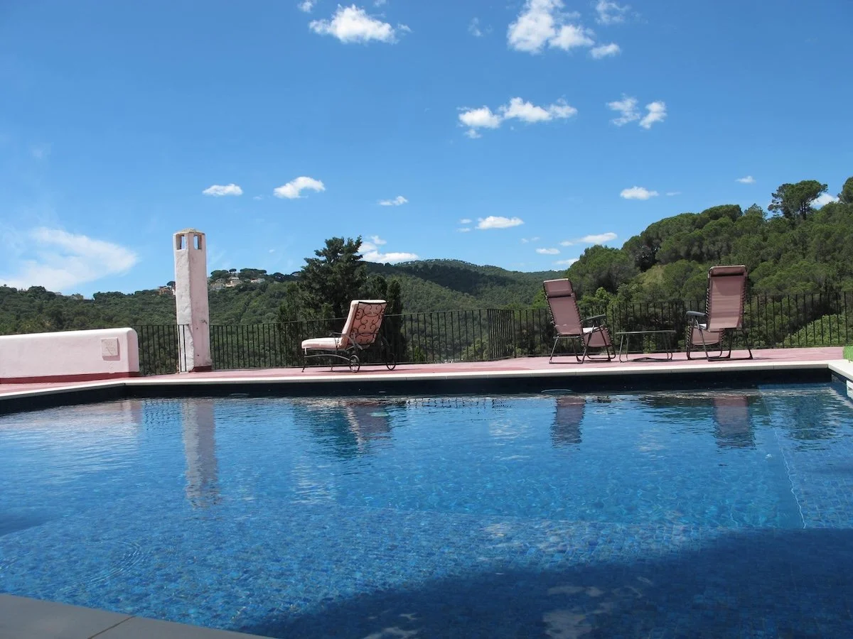 Swimming pool with lounge chairs and scenic green hills in the background under a blue sky with clouds.